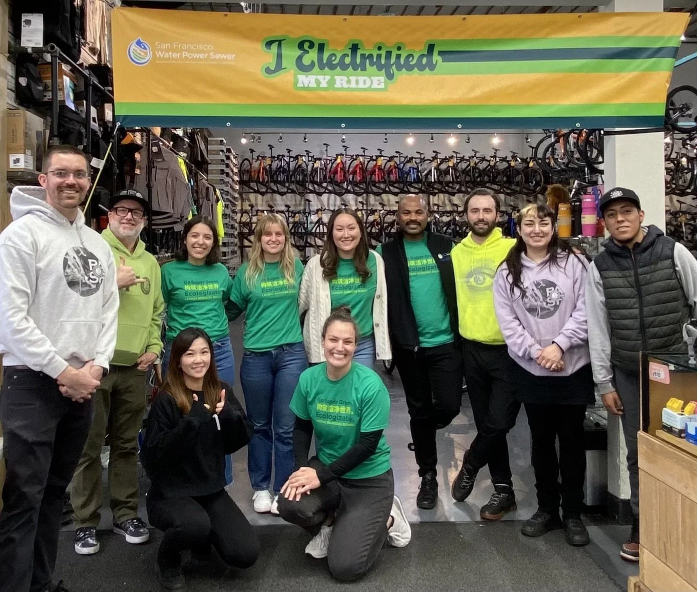 A group of eleven people, some in green I ELECTRIFIED MY RIDE shirts, pose and smile under a yellow and green banner inside a bike shop with many bicycles displayed in the background.