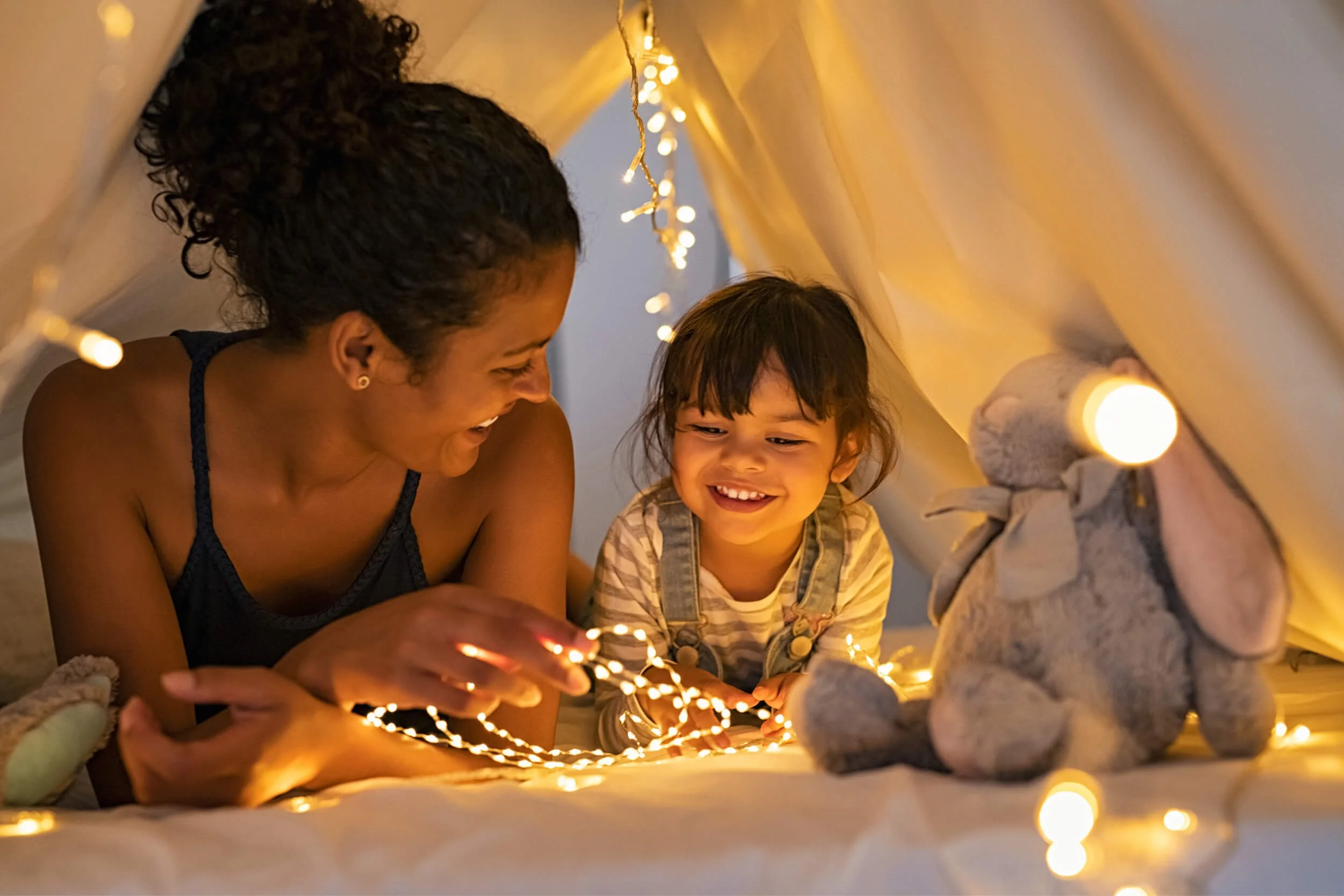 A parent and their child under their sheets and surrounded by small lights.