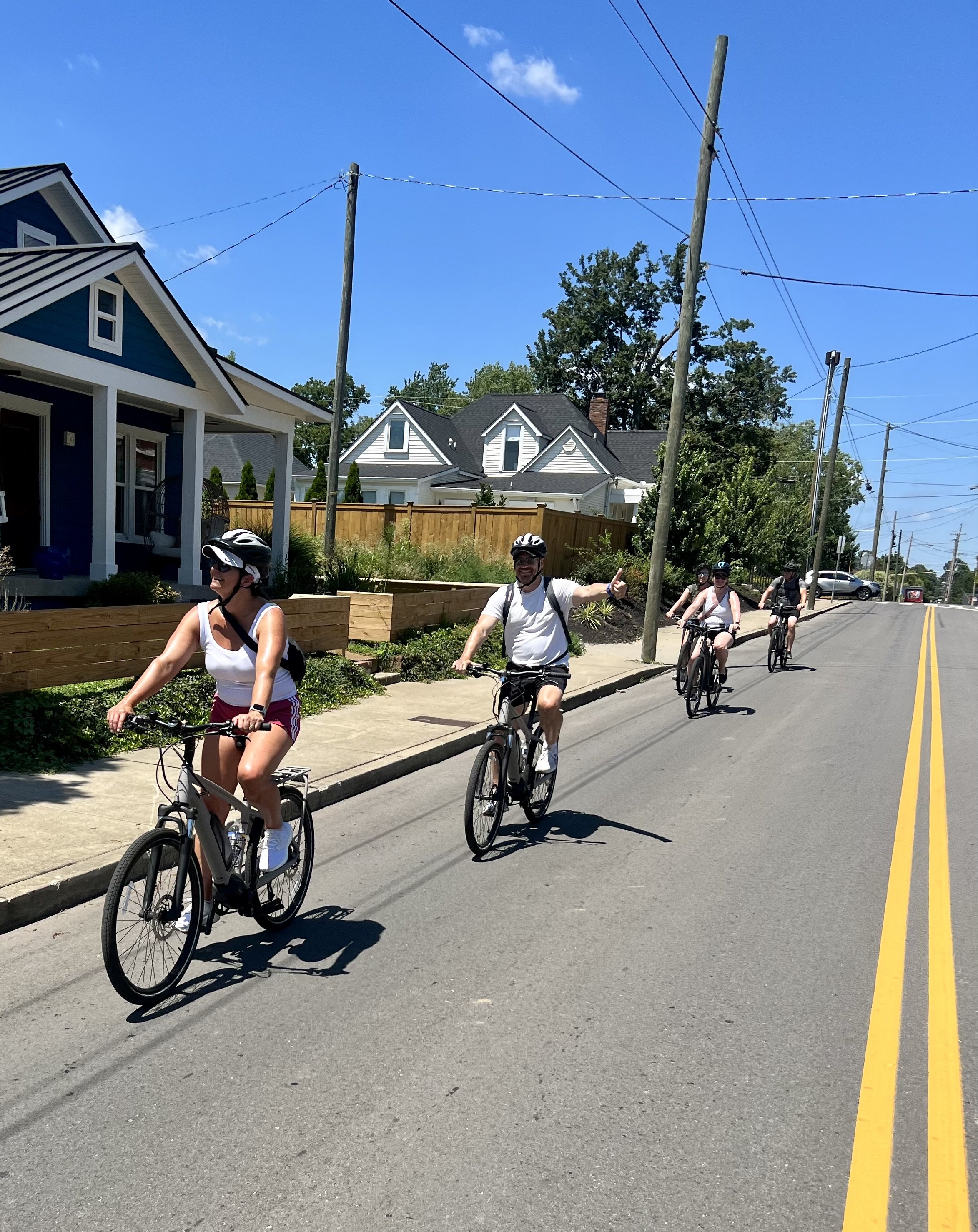 Group of four people riding bicycles on a sunny street with houses and power lines in the background.