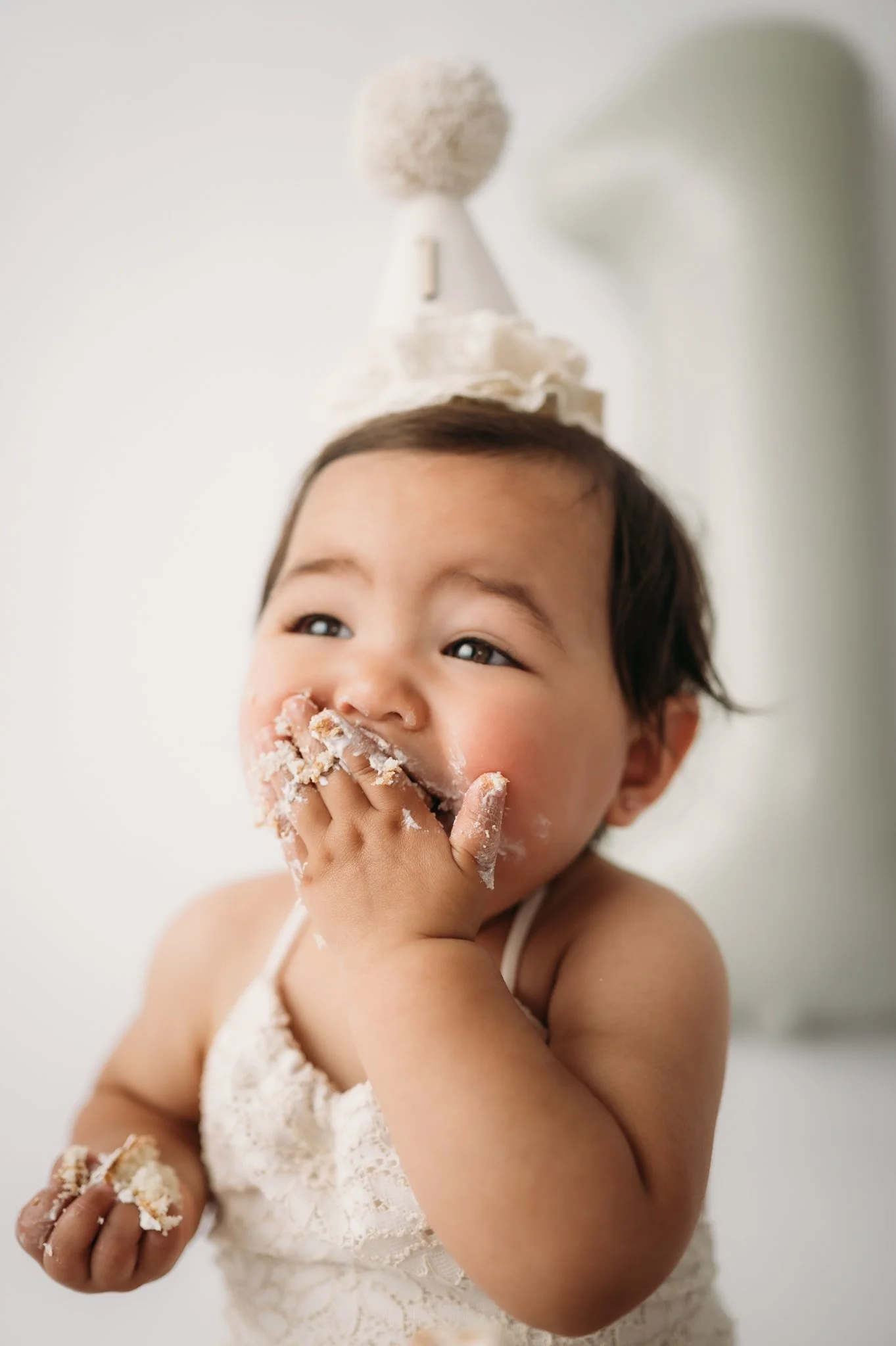 baby girl in party hat smiling with cake on hands during first birthday cake smash Houston TX