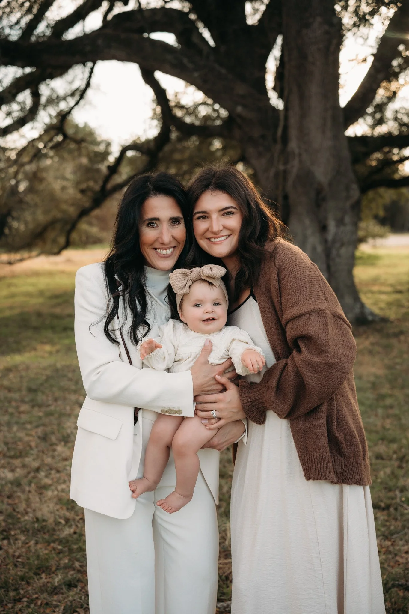 mother and grandmother smiling with baby girl under oak tree during outdoor family session The Woodlands TX