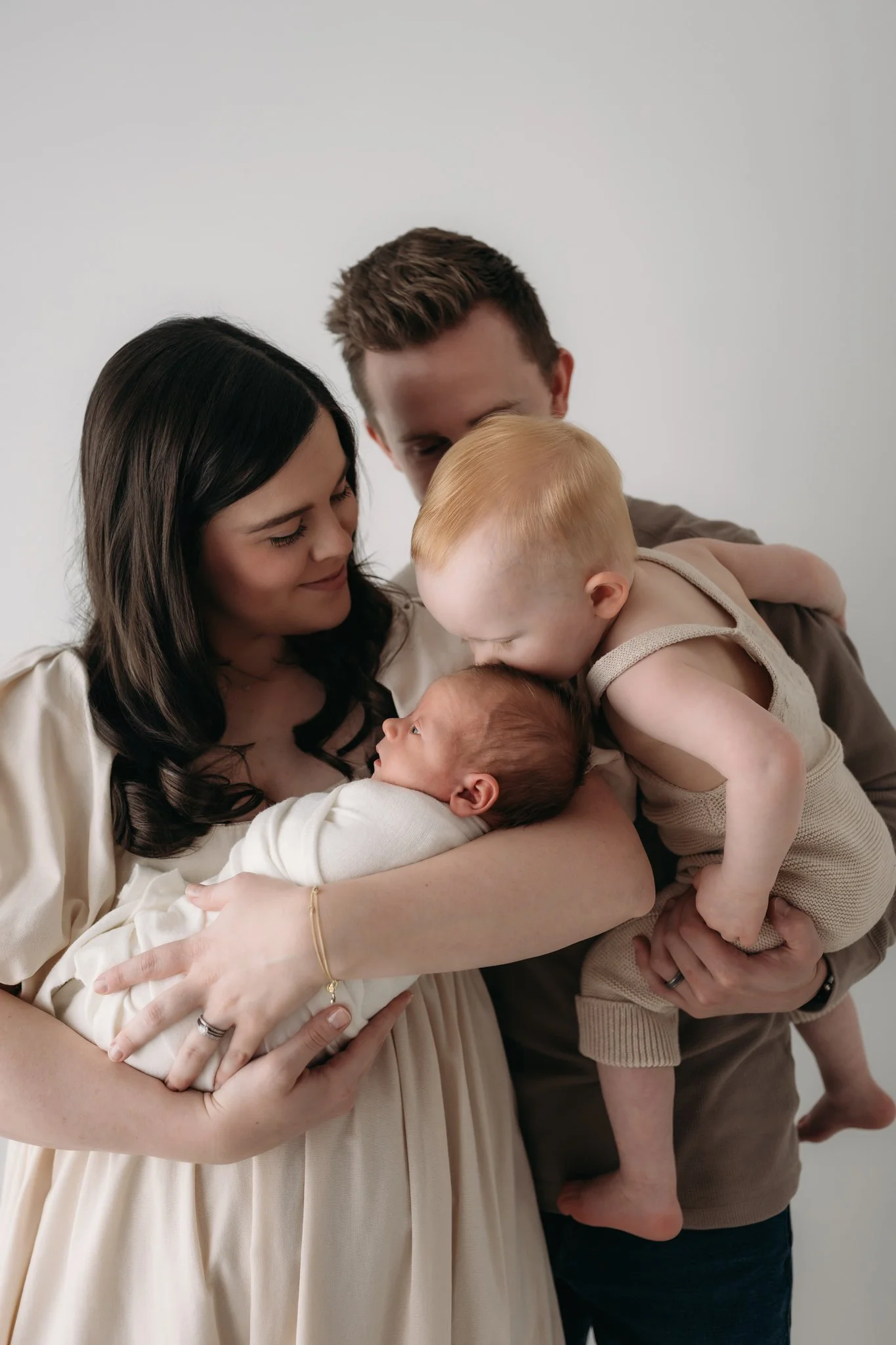parents gazing at toddler kissing sleeping newborn baby during studio newborn session The Woodlands TX