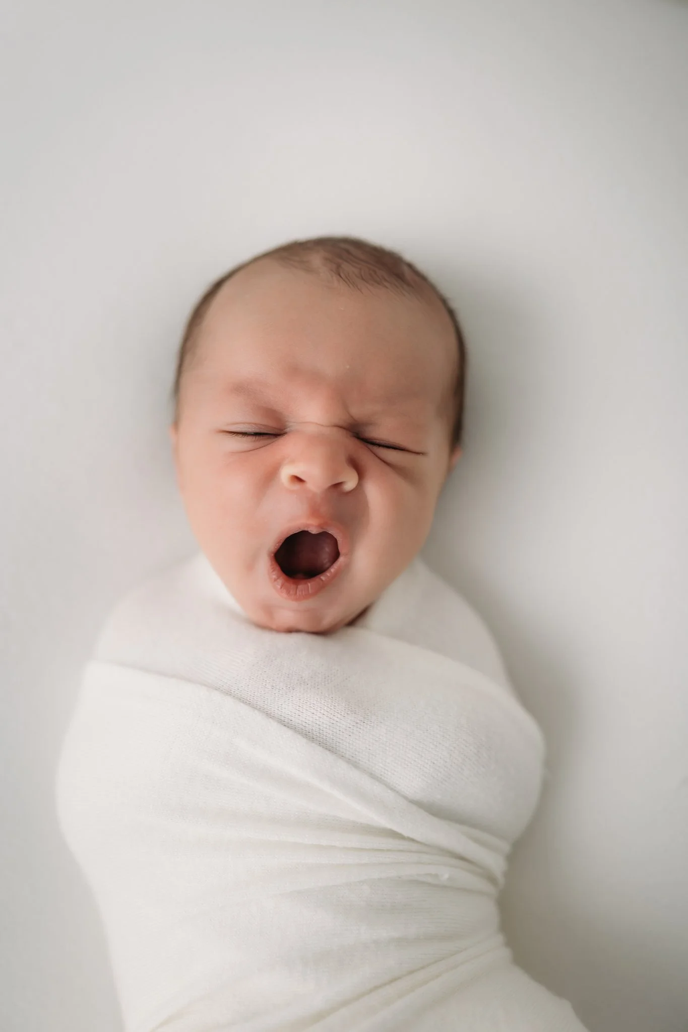 Newborn baby wrapped in white swaddle yawning during studio newborn photography session