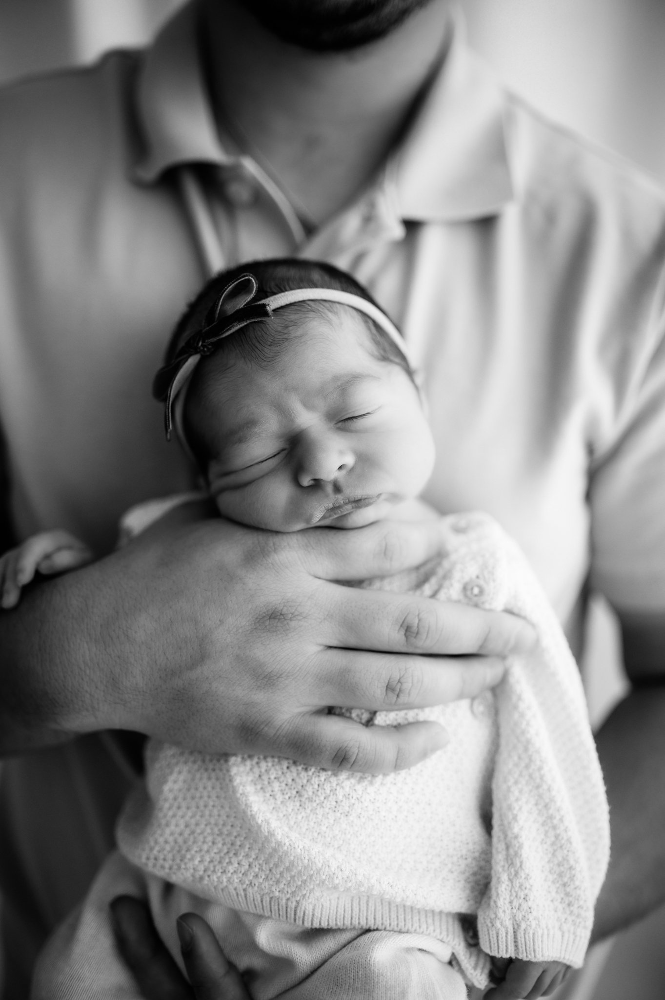 black and white close up of sleeping newborn baby girl with bow headband cradled in parent's hands Houston TX