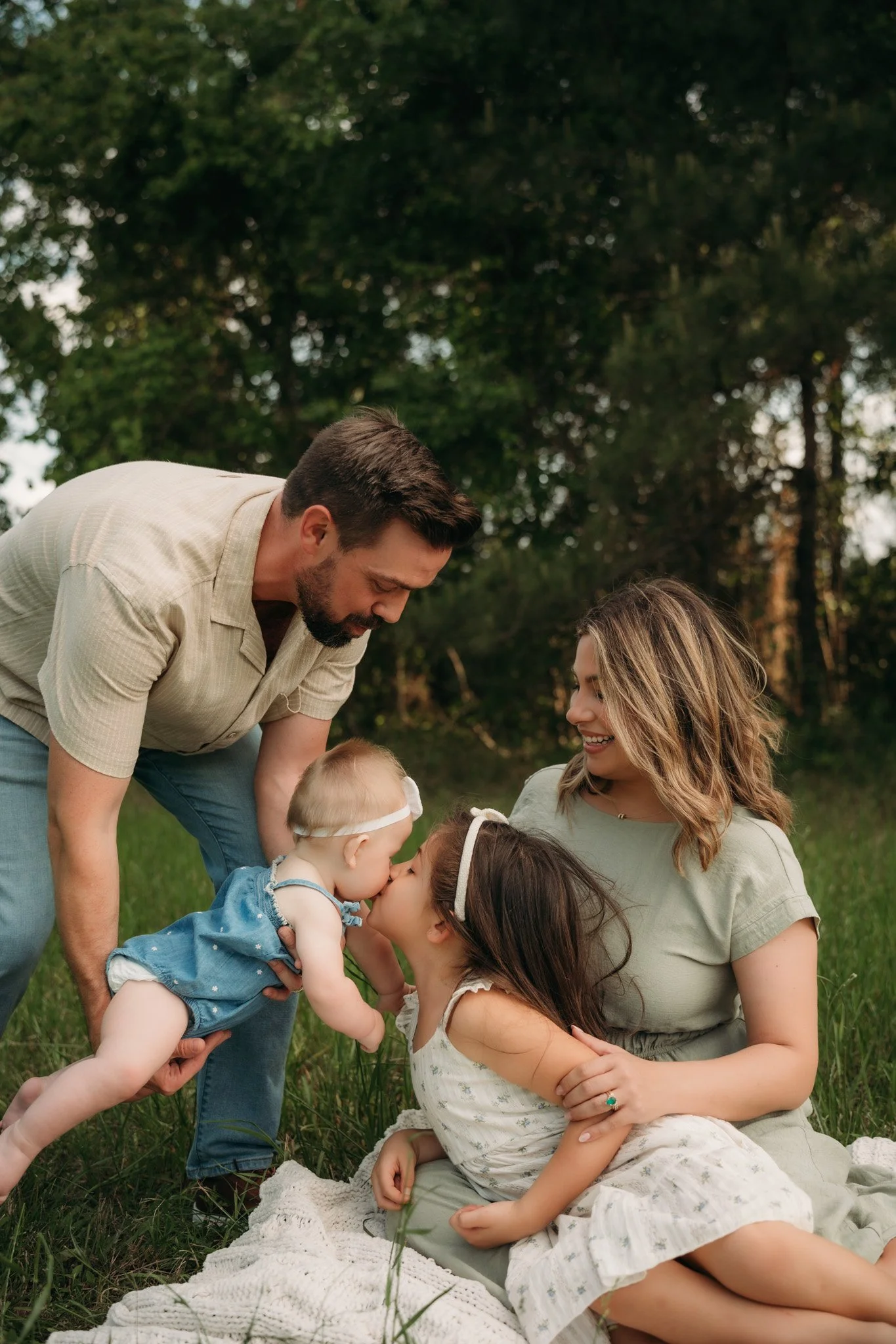 Outdoor family session in Houston, Texas with parents and two young daughters sharing a sweet kiss in a grassy field surrounded by trees at golden hour.