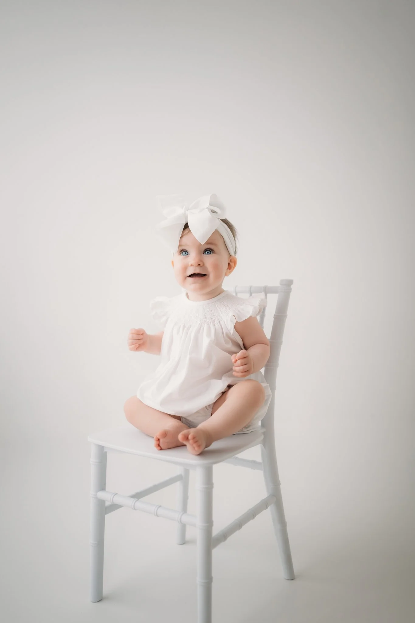 Baby girl in white dress and bow sitting on a chair during a light and airy milestone photography session in Houston, Texas