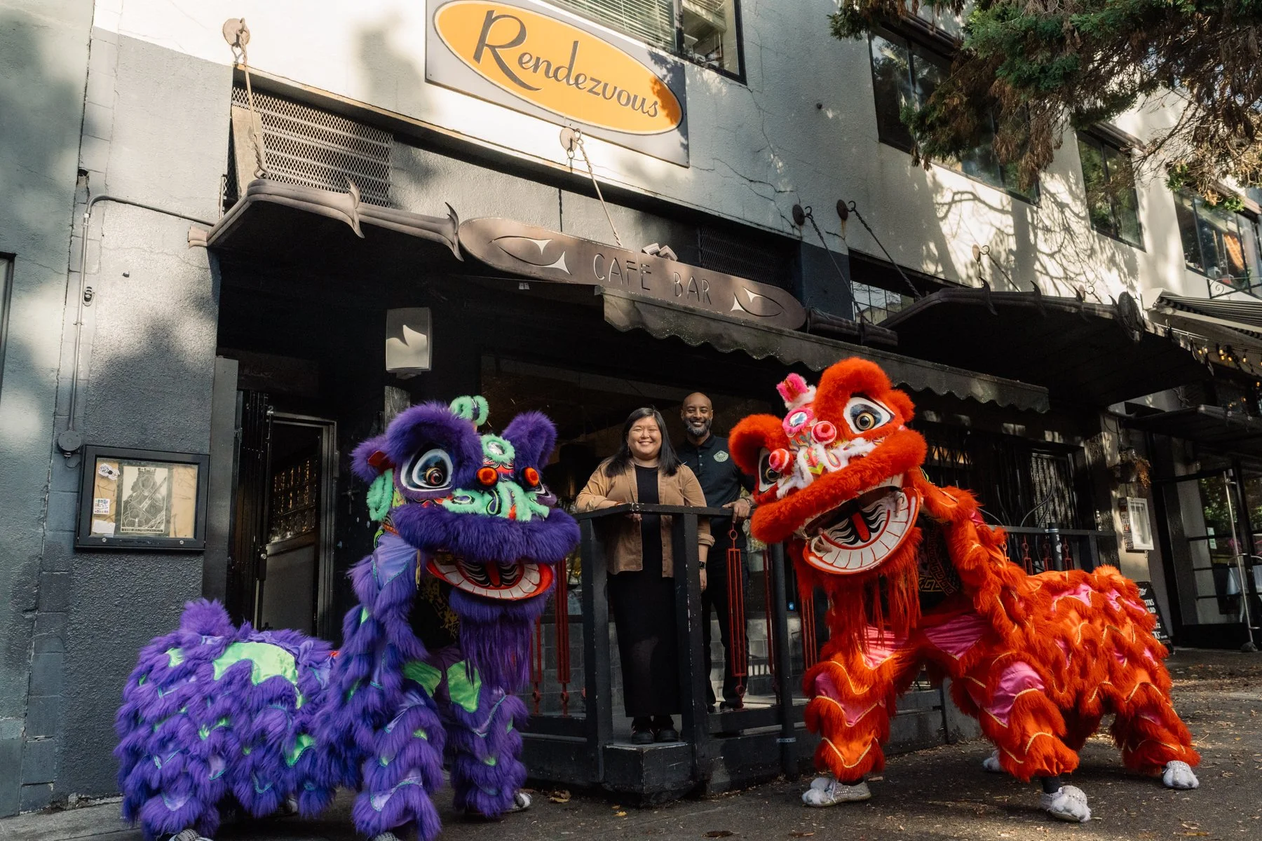 Two people stand on a small black balcony with two colorful lion dance costumes flanking them, one purple and one red, in front of a gray building with the sign 'Rendezvous' and another sign reading 'Cafe Bar.'