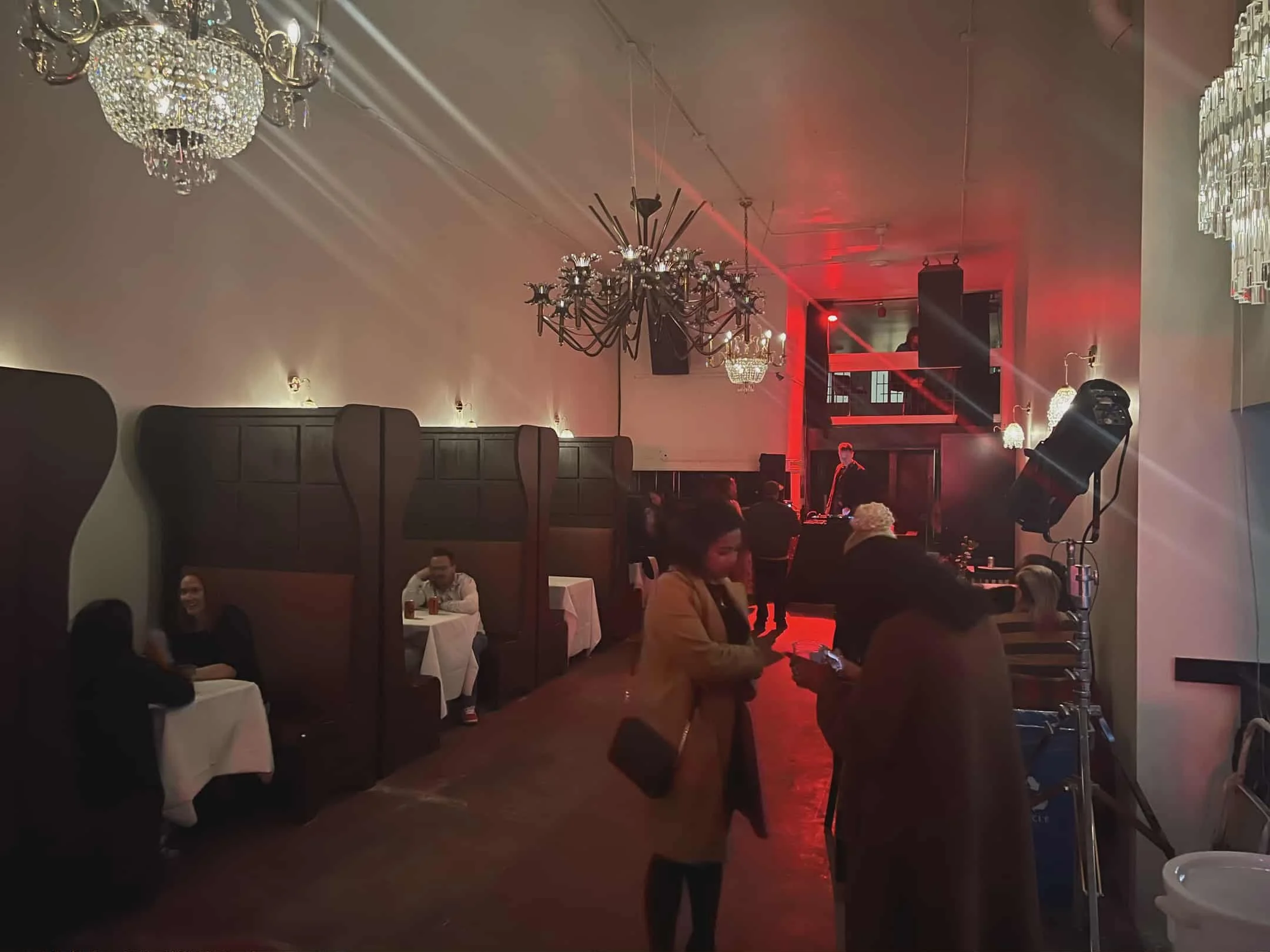 A dimly lit event space with chandeliers and wall sconces. Several people are seated at tables with white tablecloths, some of whom are talking or looking at their phones. In the foreground, two women are having a conversation, with one looking at her phone. Toward the back of the room, a DJ or performer is visible near a DJ booth, with red lighting and fog effects adding to the ambiance.