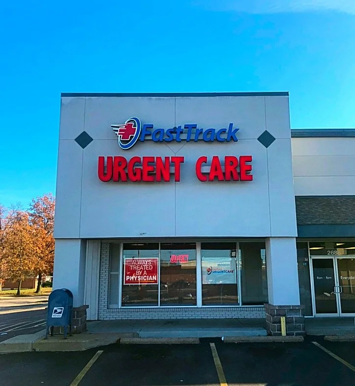 Facade of FastTrack Urgent Care clinic with a large sign, glass entrance door, and a USPS mailbox outside, under a clear blue sky.
