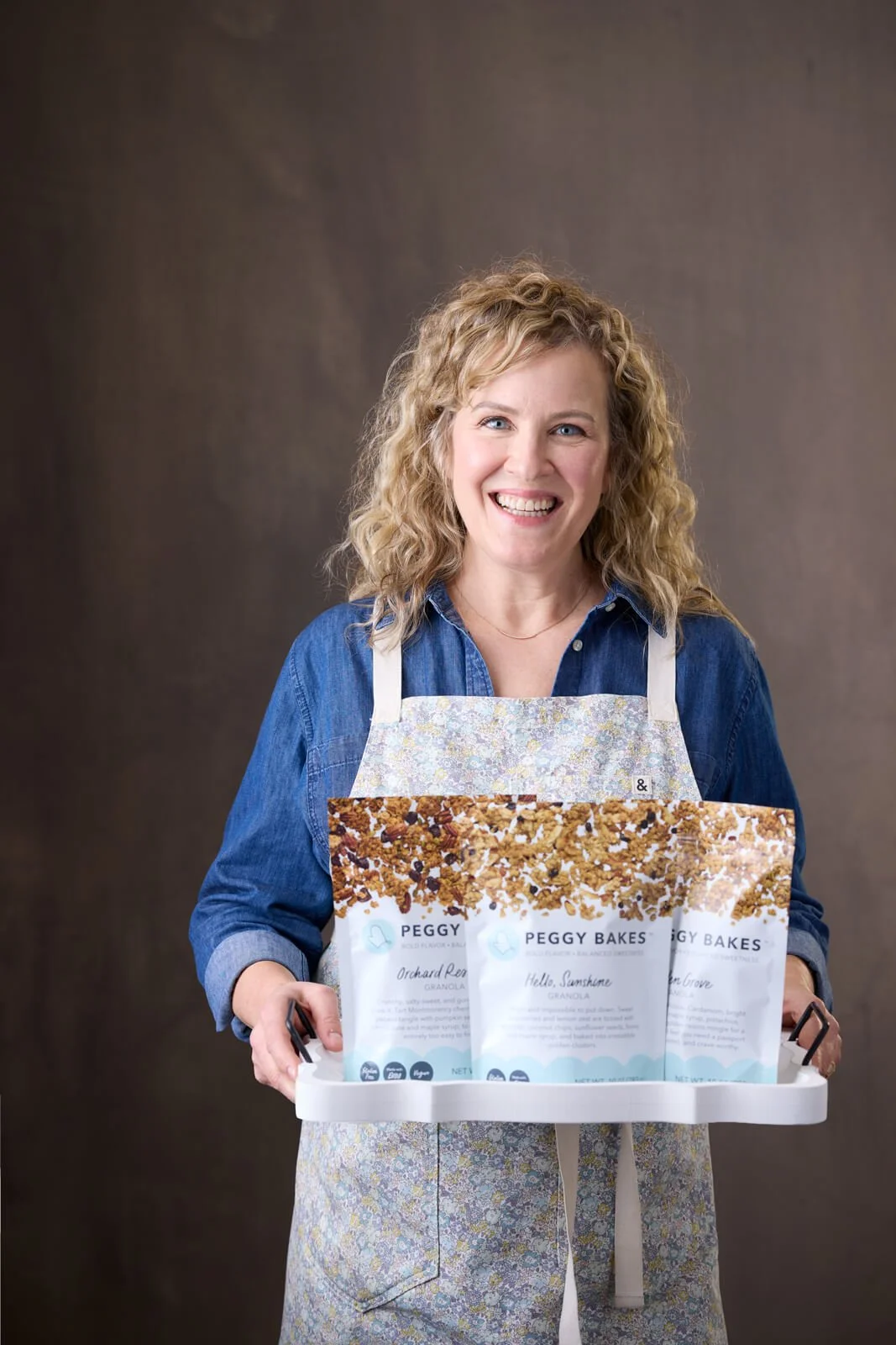 Peggy Bakes founder, Peggy Sonnenburg, holding a tray of her granola. Includes bags of Hello, Sunshine, Golden Grove and Orchard Reserve