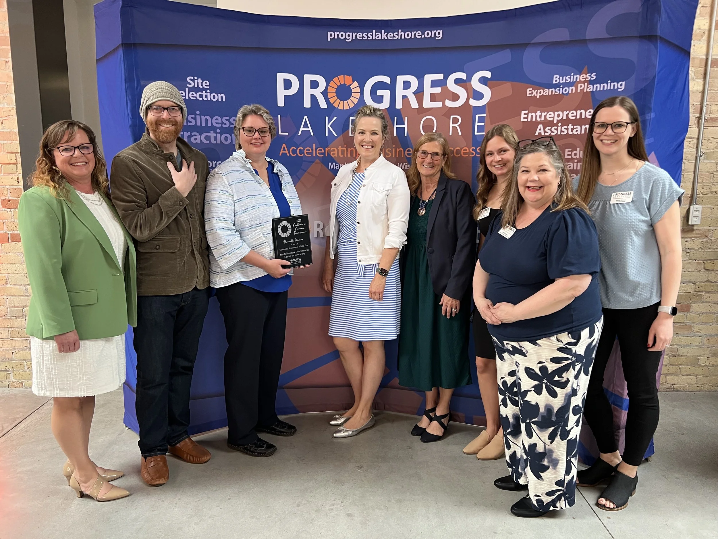 Peggy Bakes founder, Peggy Sonnenburg standing infant of a Progress Lakeshore banner with a group of people