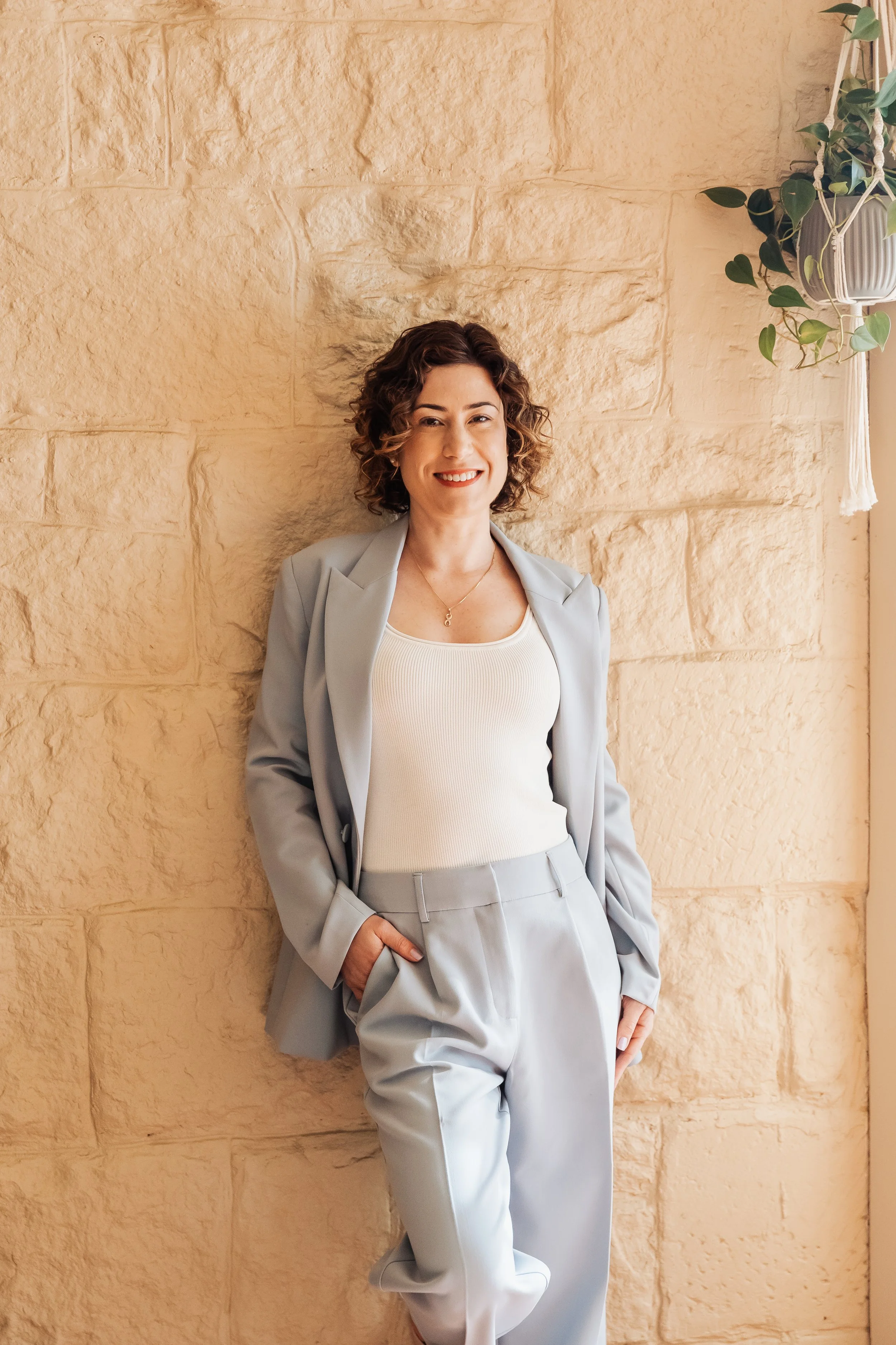 Jaclyn Hall PACFA Accredited Clinical Counsellor and Certified Supervisor, in a light gray blue suit with a white top and a necklace is smiling and posing against a beige stone wall with a hanging plant on the right side.