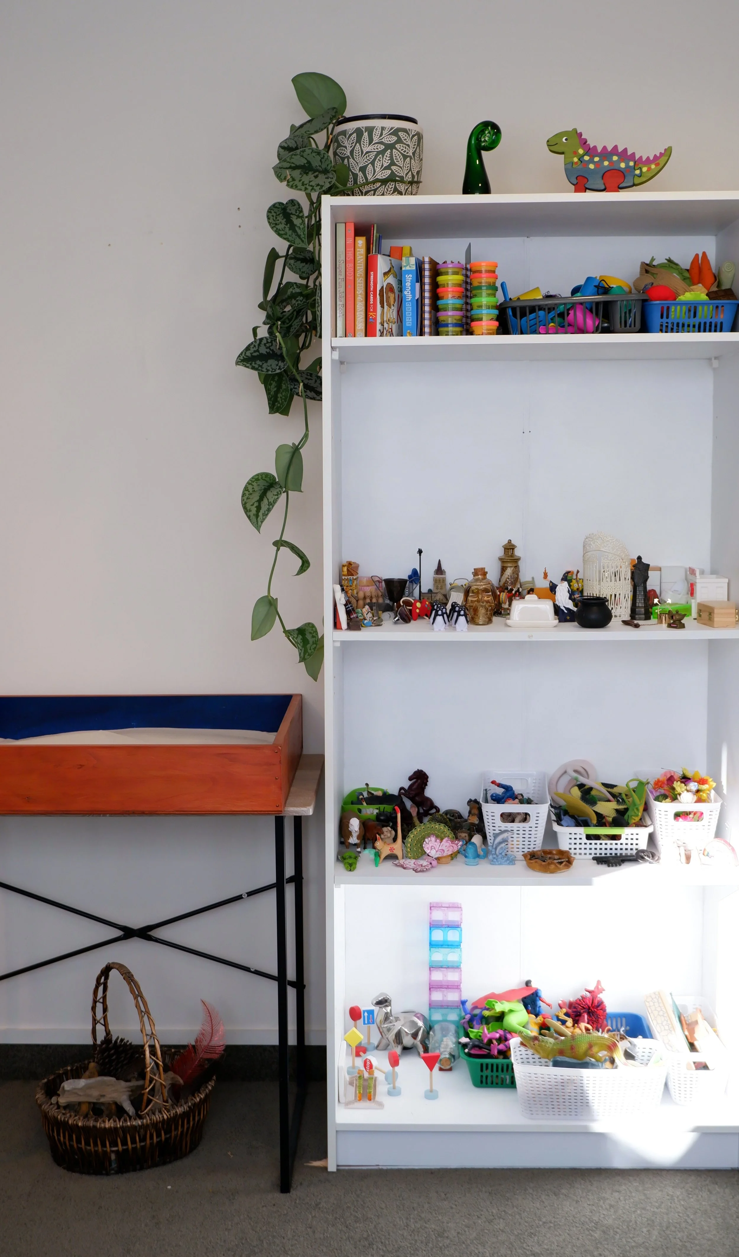 A white shelf filled with colorful toys, books, and decorative items, with a green leafy houseplant hanging on a wall next to it. To the left is a table with a wooden tray, and a basket with a woven toy underneath.