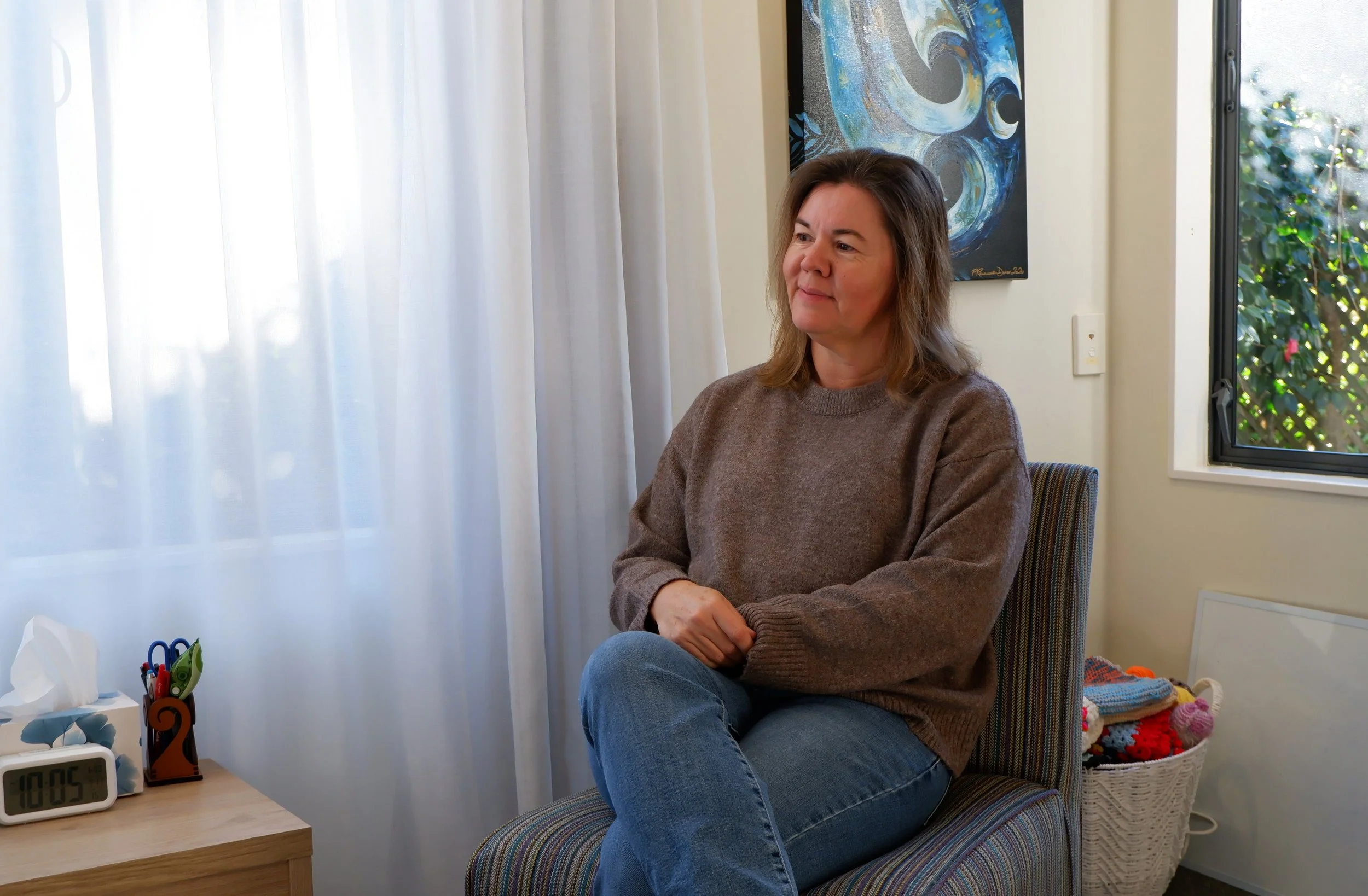 A woman sitting in a room with sunlight coming through white curtains, with a colorful abstract painting on the wall behind her. She is wearing a brown sweater and blue jeans, with her hands resting on her lap. There is a night clock showing the time 18:05 on a wooden table alongside a tissue box, pens in a holder, and a basket of crocheted items.
