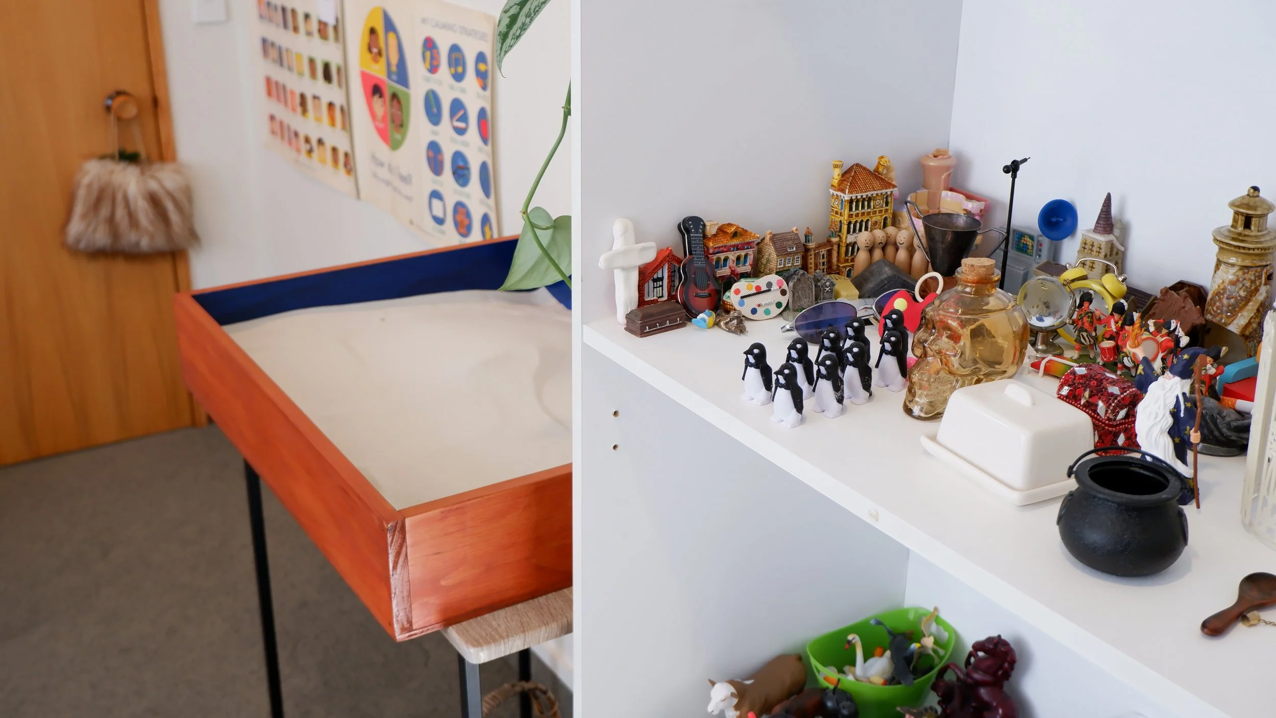 Display of various small decorative and toy items on a white shelf and a nearby table, including miniature buildings, figurines, a guitar, a skull, a lamp, and other assorted objects.