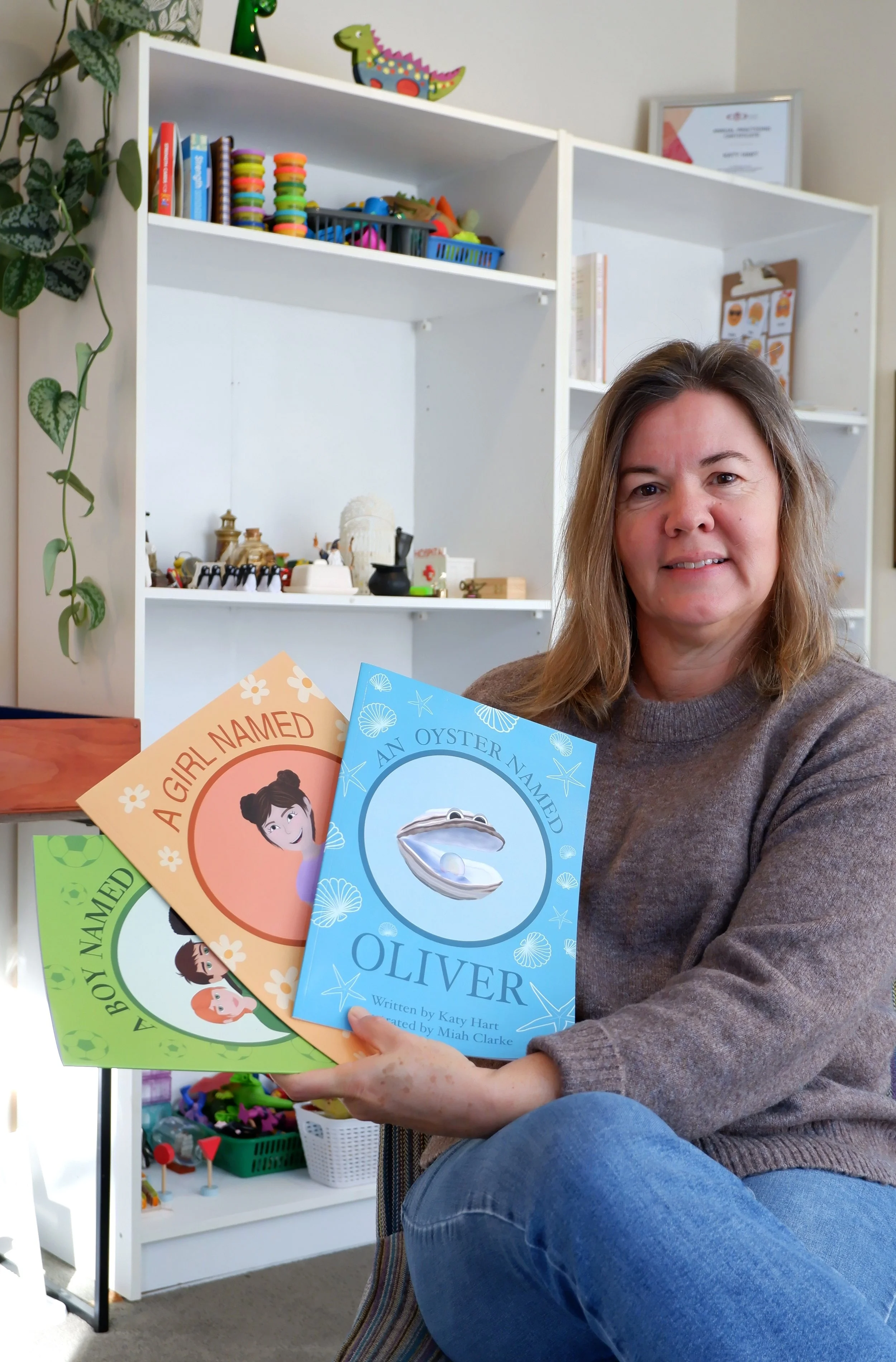 Woman sitting on a chair holding three children's books titled 'A Girl Named Olivia', 'An Oyster Named Olivia', and 'A Boy Named Oliver'. Behind her is a white shelf filled with toys, books, and decorative items. A plant hangs on the left side of the shelf.