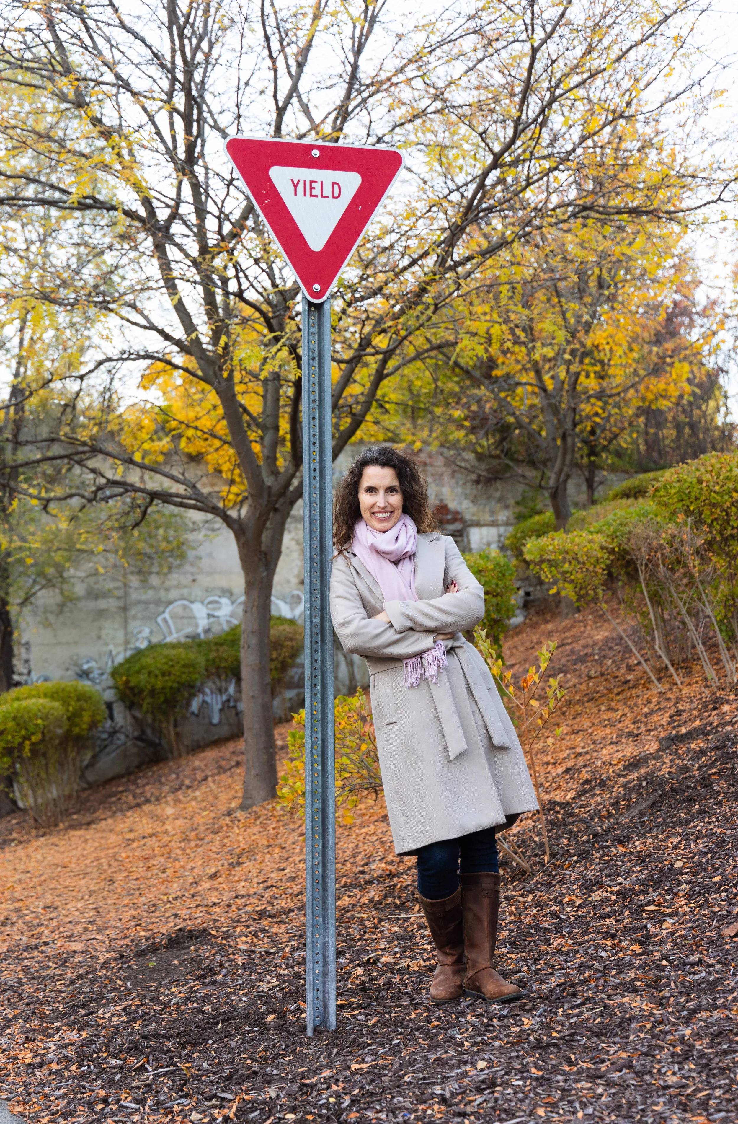 A person in a beige coat and pink scarf leaning on a yield sign in a park with autumn trees.