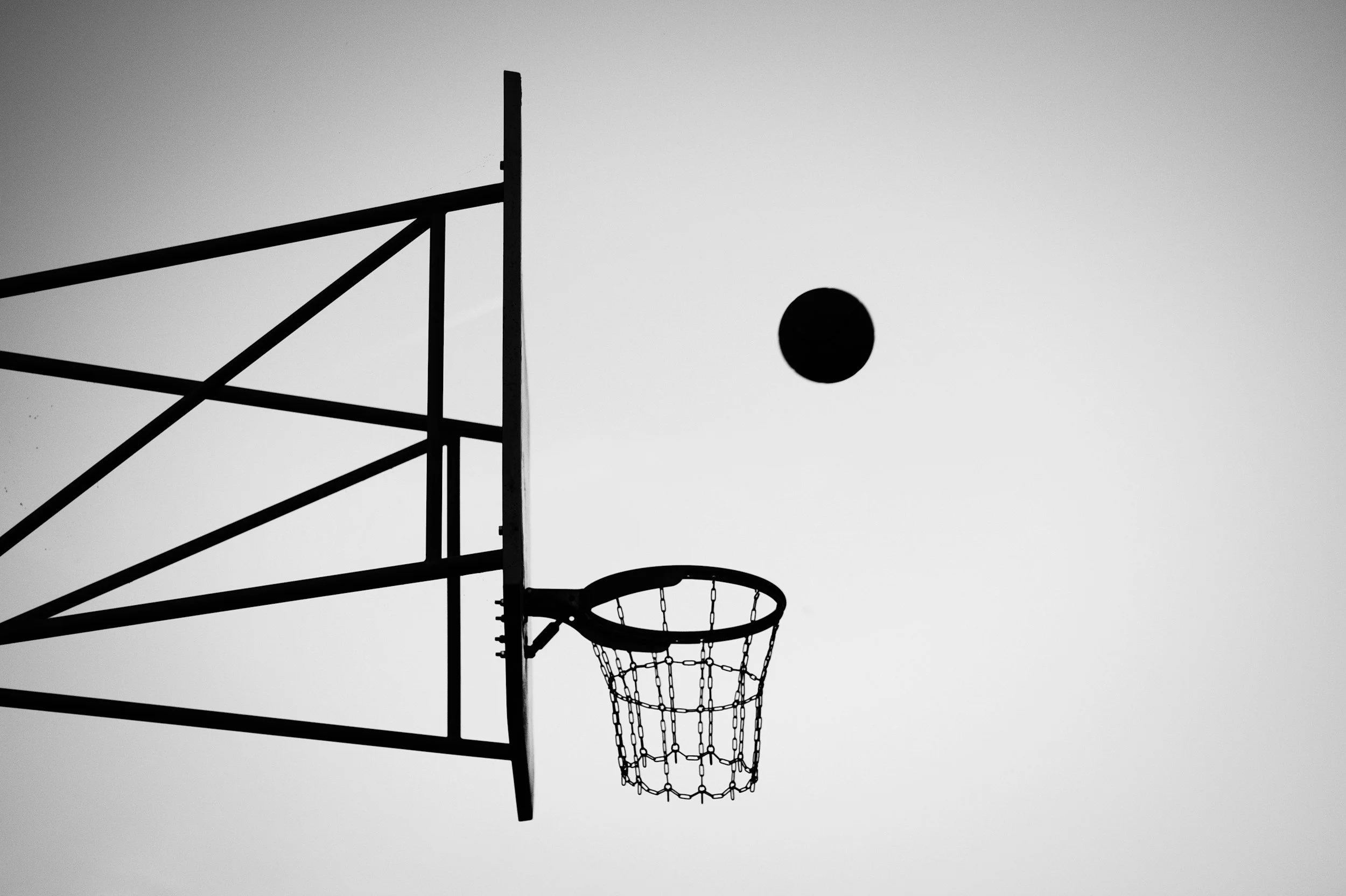 Black and white silhouette of a basketball hoop and backboard with a basketball in the air, against a clear sky.