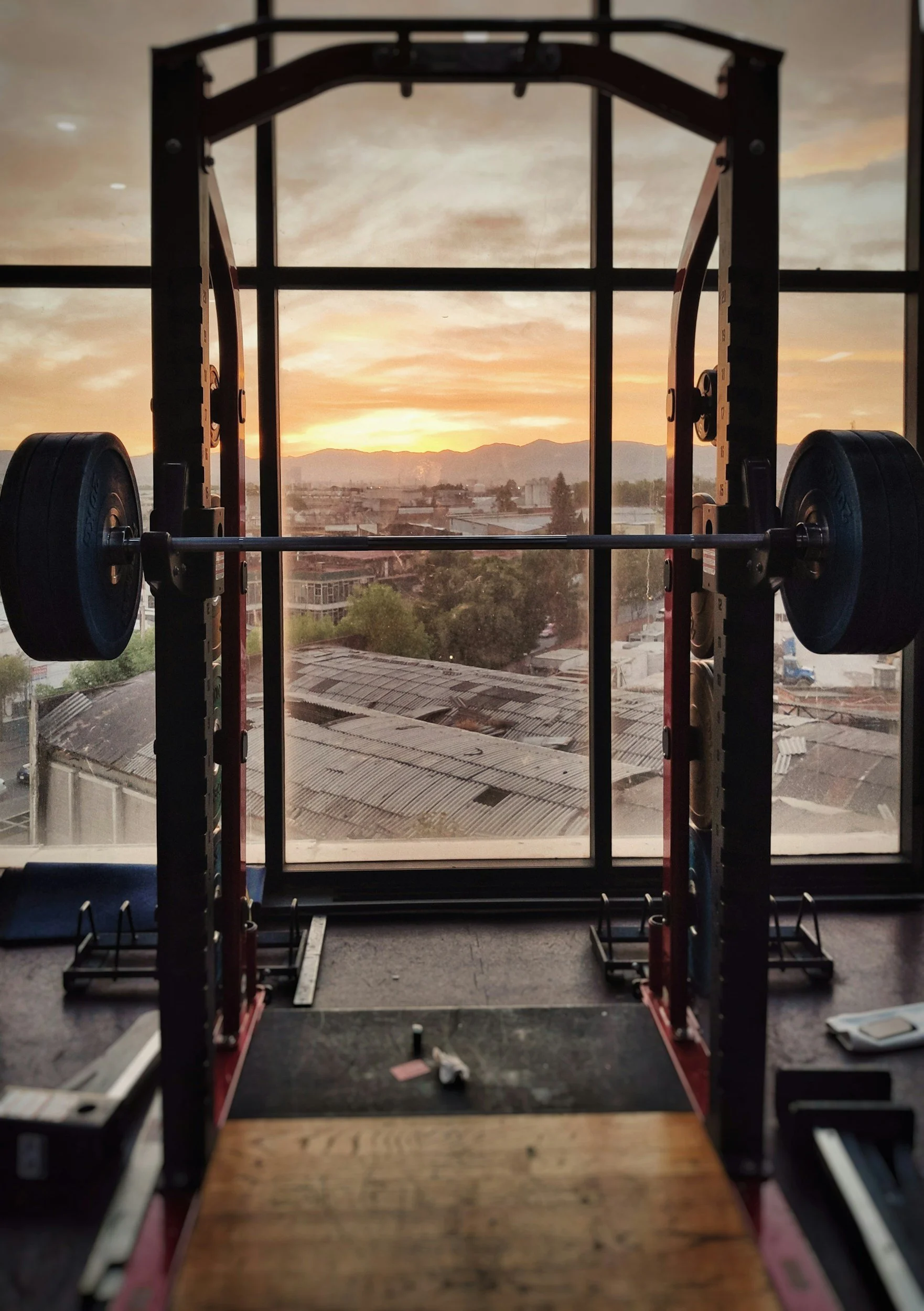 A squat rack with a barbell and weight plates in a gym, set against a large window showing a cityscape at sunset.