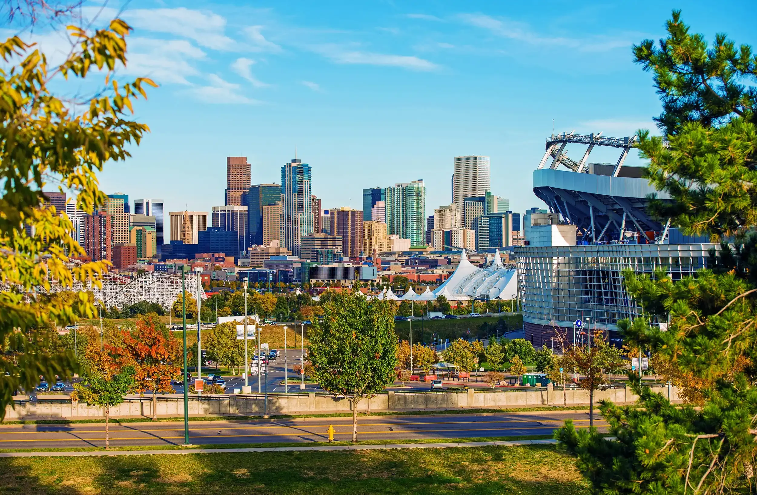 A city skyline with tall buildings, a stadium structure on the right, trees in the foreground, and a partly cloudy blue sky.