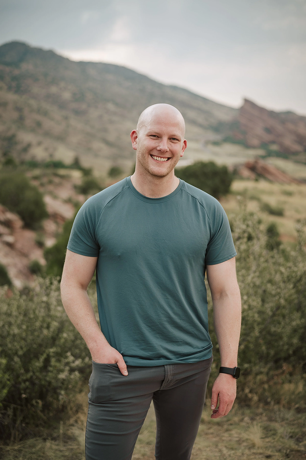 Smiling man with a bald head wearing a teal athletic shirt and dark pants, standing outdoors in a scenic landscape with mountains, sparse trees, and cloudy sky in the background.