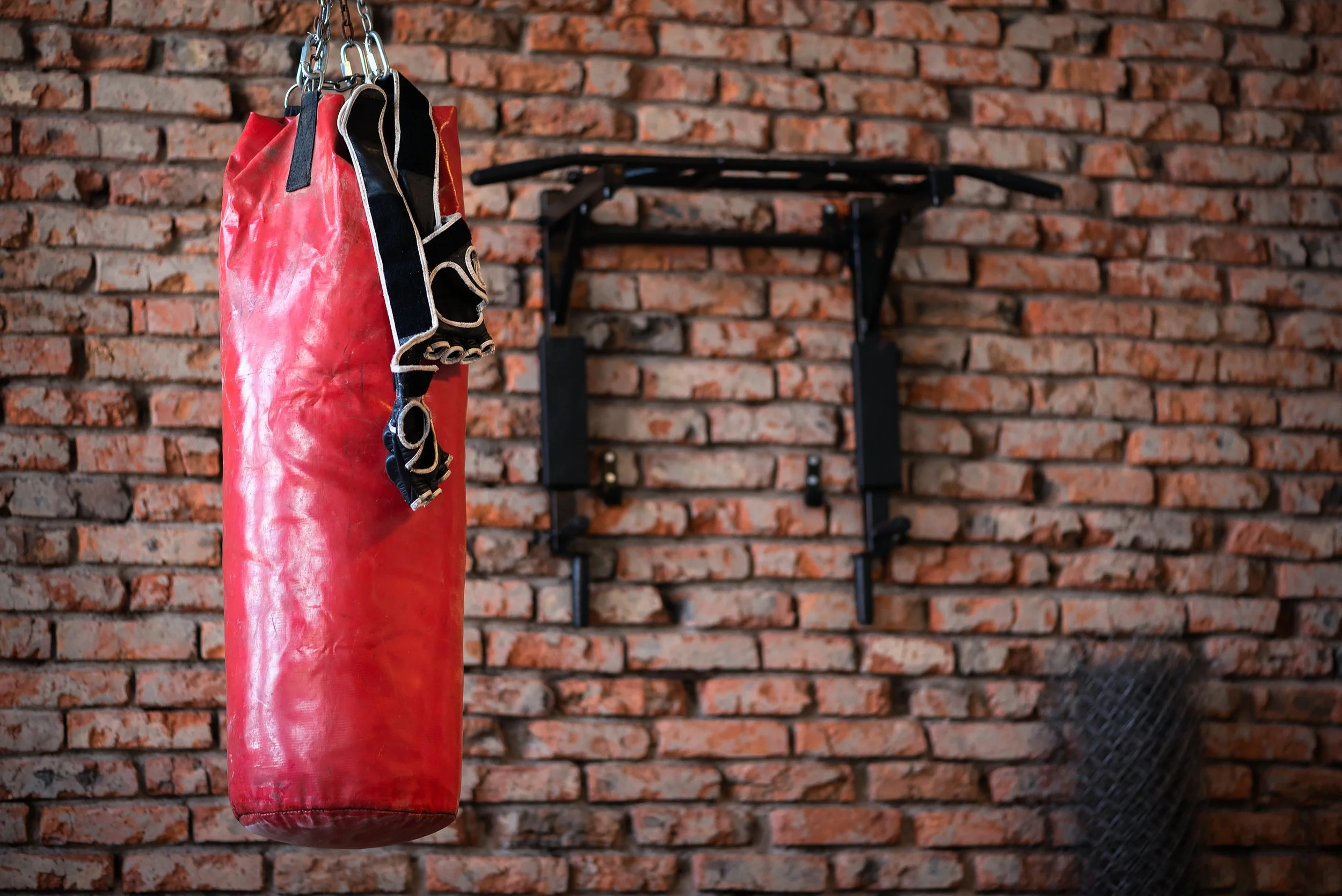 A red punching bag hanging in front of a brick wall with boxing gloves draped over it.