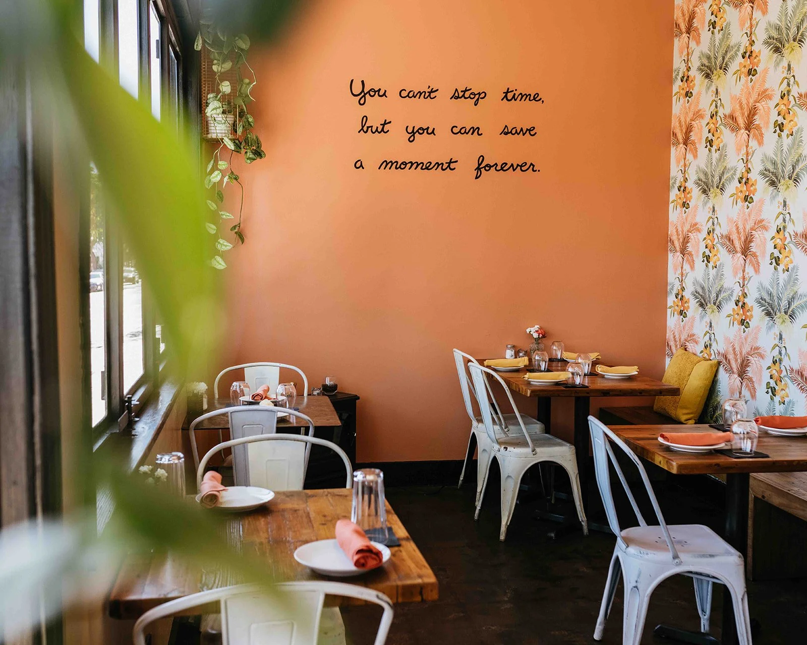A cozy restaurant interior with wooden tables, white chairs, and pastel decor. A peach-colored wall features a quote: 'You can't stop time, but you can save a moment forever.' There's a patterned wallpaper with palm leaves and flowers on one wall, and green plants near the window.