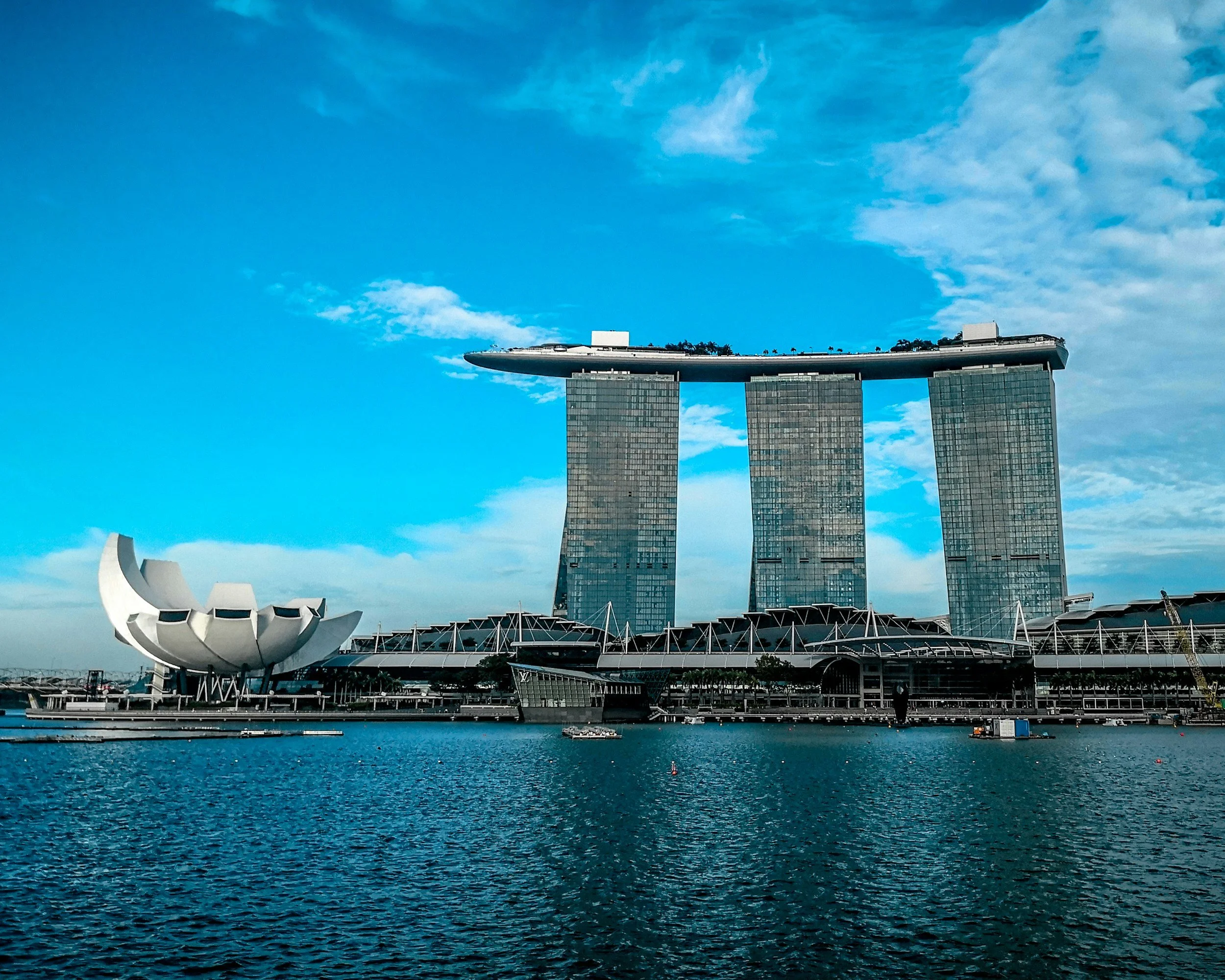 Photo of the Marina Bay Sands hotel in Singapore, with three tall glass towers and a boat-shaped rooftop structure, along with the ArtScience Museum shaped like a lotus flower on the left, and water in the foreground under a blue sky with clouds.