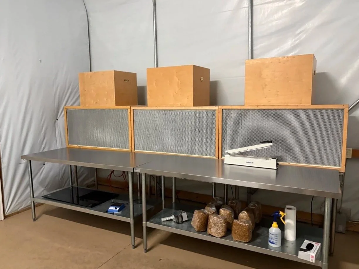 A stainless steel table with three large wooden boxes on top, three wooden soundproof panels behind the boxes, and a small white device on a long metal counter. There are rocks, a spray bottle, paper towels, and some tools underneath the table.