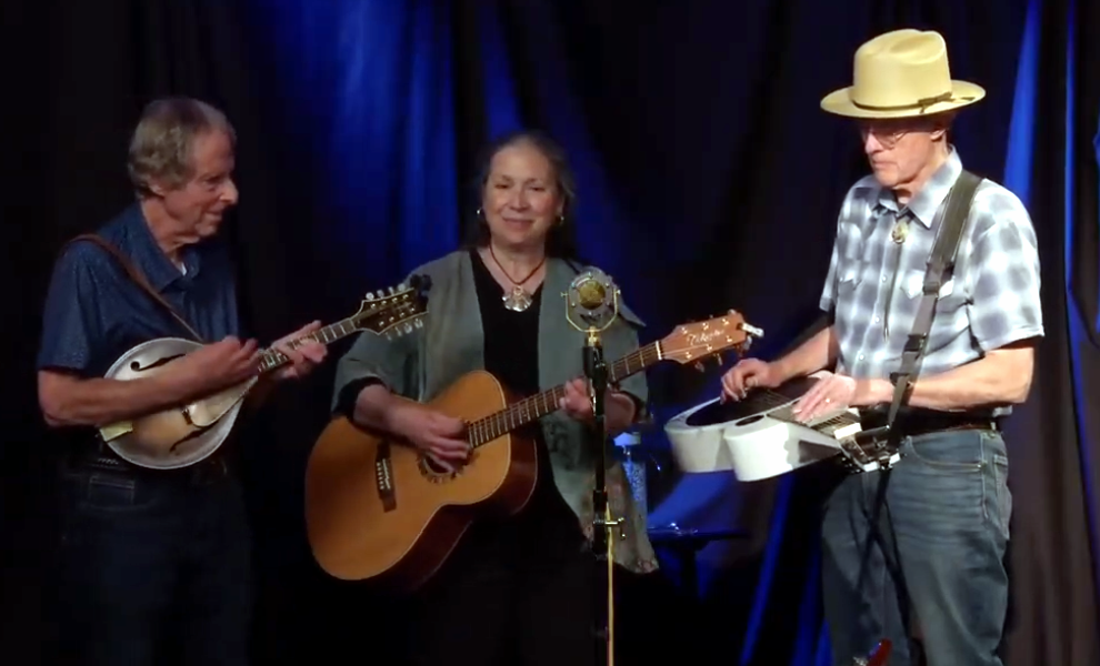 Three musicians performing on stage, playing string instruments and a synthesizer, with a woman singing into a microphone, in front of a dark blue curtain.