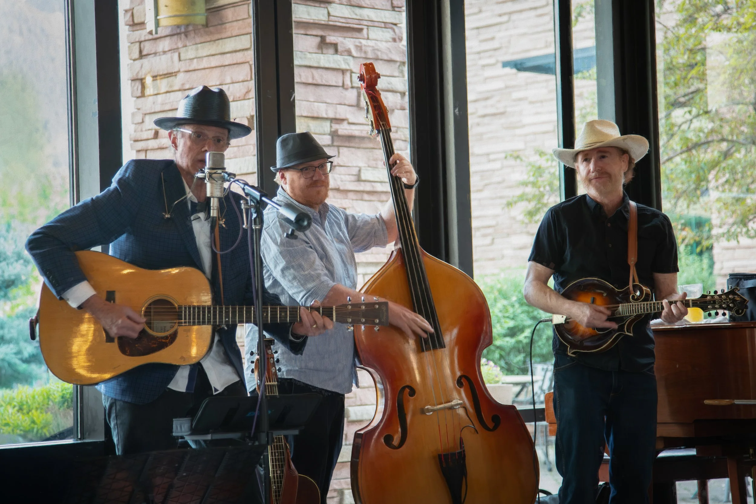 Three musicians performing indoors with large windows behind them. The man on the left is playing an acoustic guitar and singing into a microphone, wearing a dark plaid jacket and a black hat. The man in the middle is playing an upright bass, wearing glasses, a striped shirt, and a gray fedora. The man on the right is playing a mandolin, wearing a cowboy hat and a black shirt. Outside, trees and a building are visible through the windows.