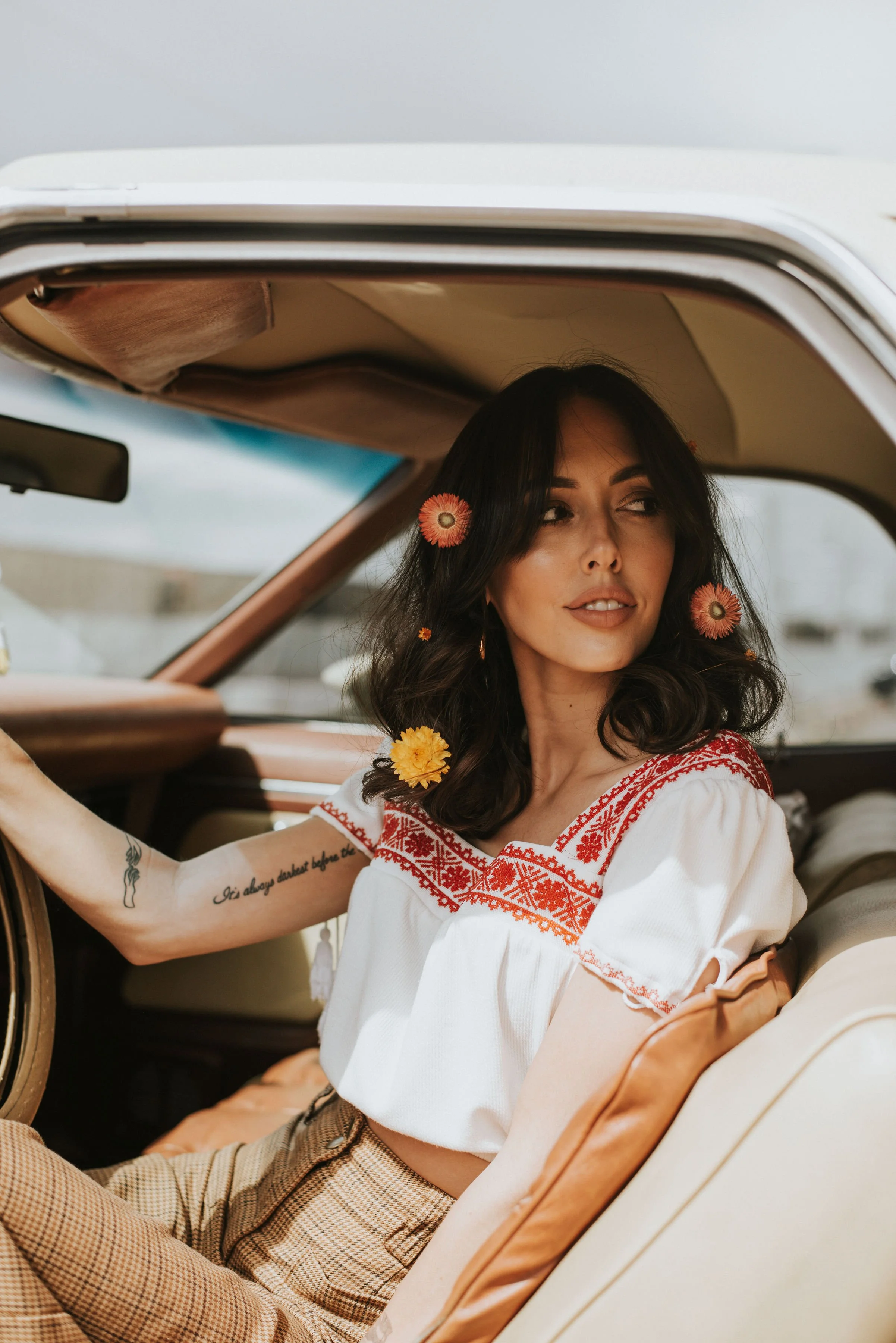 A woman sits in a vintage car with curtain bangs and flowers in her hair