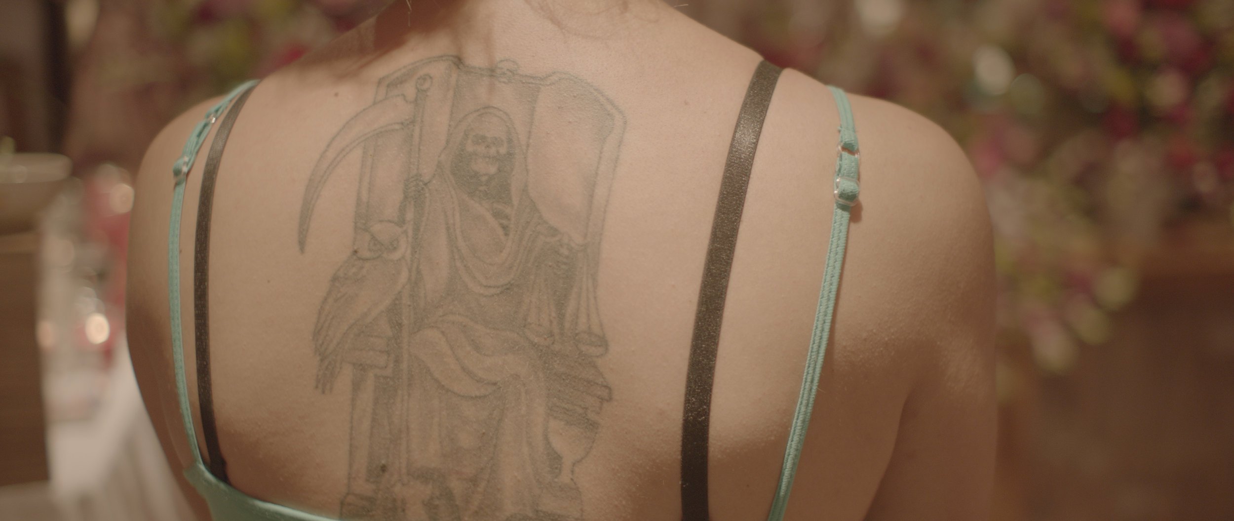 Back view of a person with a large tattoo of a Grim Reaper holding a scythe, wearing a hoodie, visible on their upper back and shoulder.
