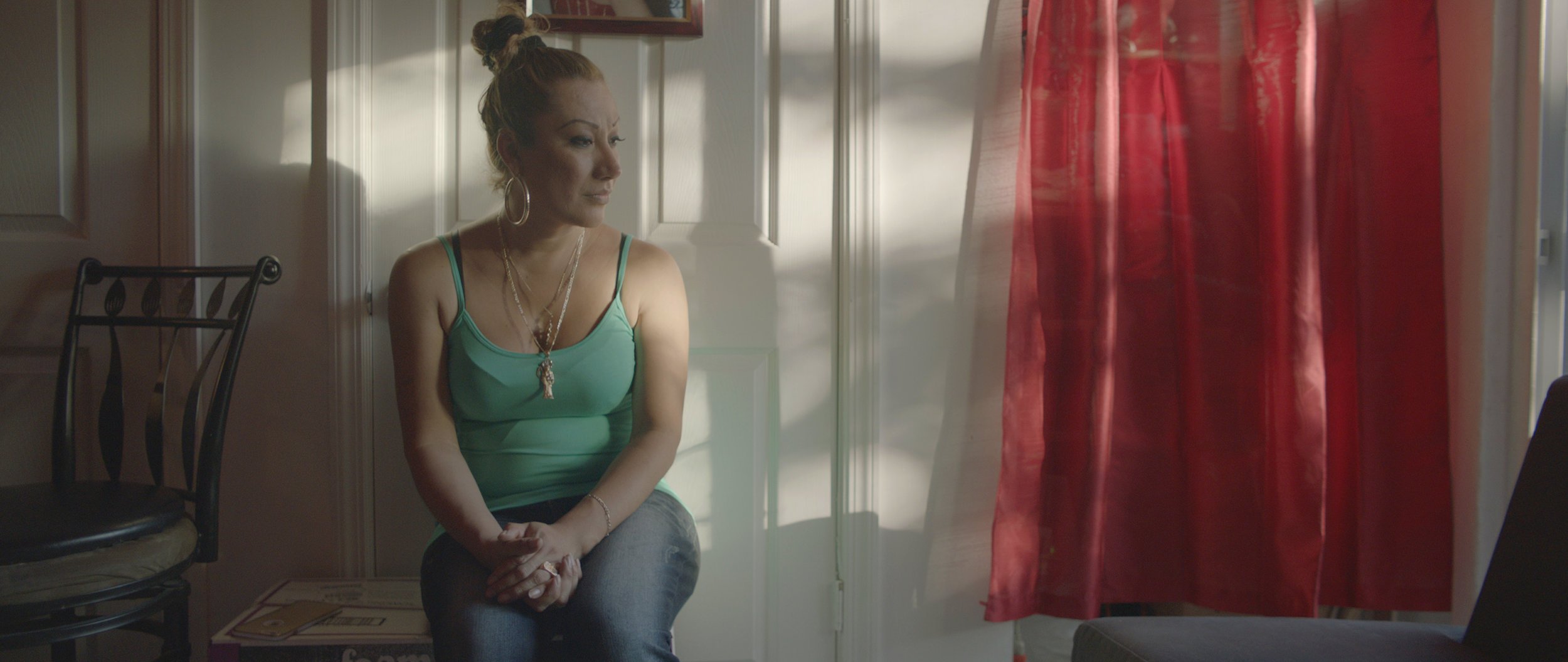 A woman with blonde hair tied in a bun, wearing a green tank top and jewelry, sitting by a window with red curtains, looking thoughtful.