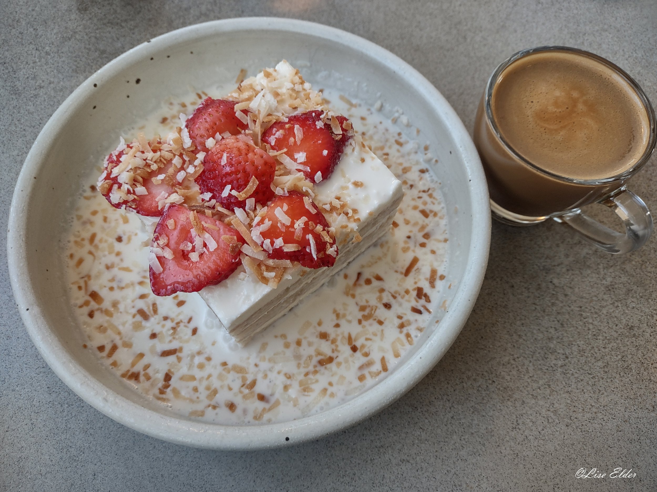 Vegan vanilla and strawberry cake in vegan cream sauce with toasted coconut flakes in a white bowl next to a glass cup of coffee.
