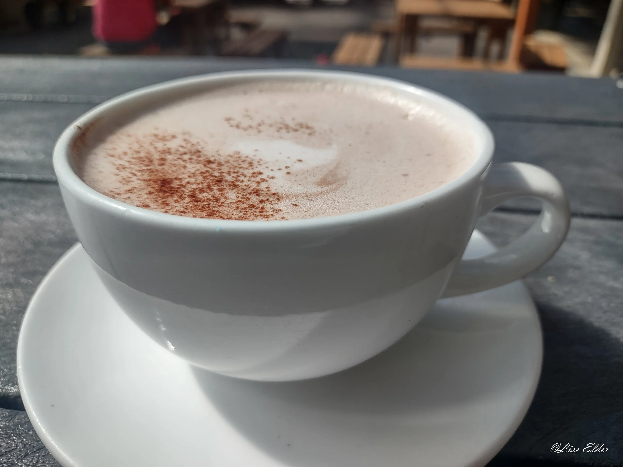 A large white cup of almond milk mocha  and cacao powder on top, at a gray wooden table.