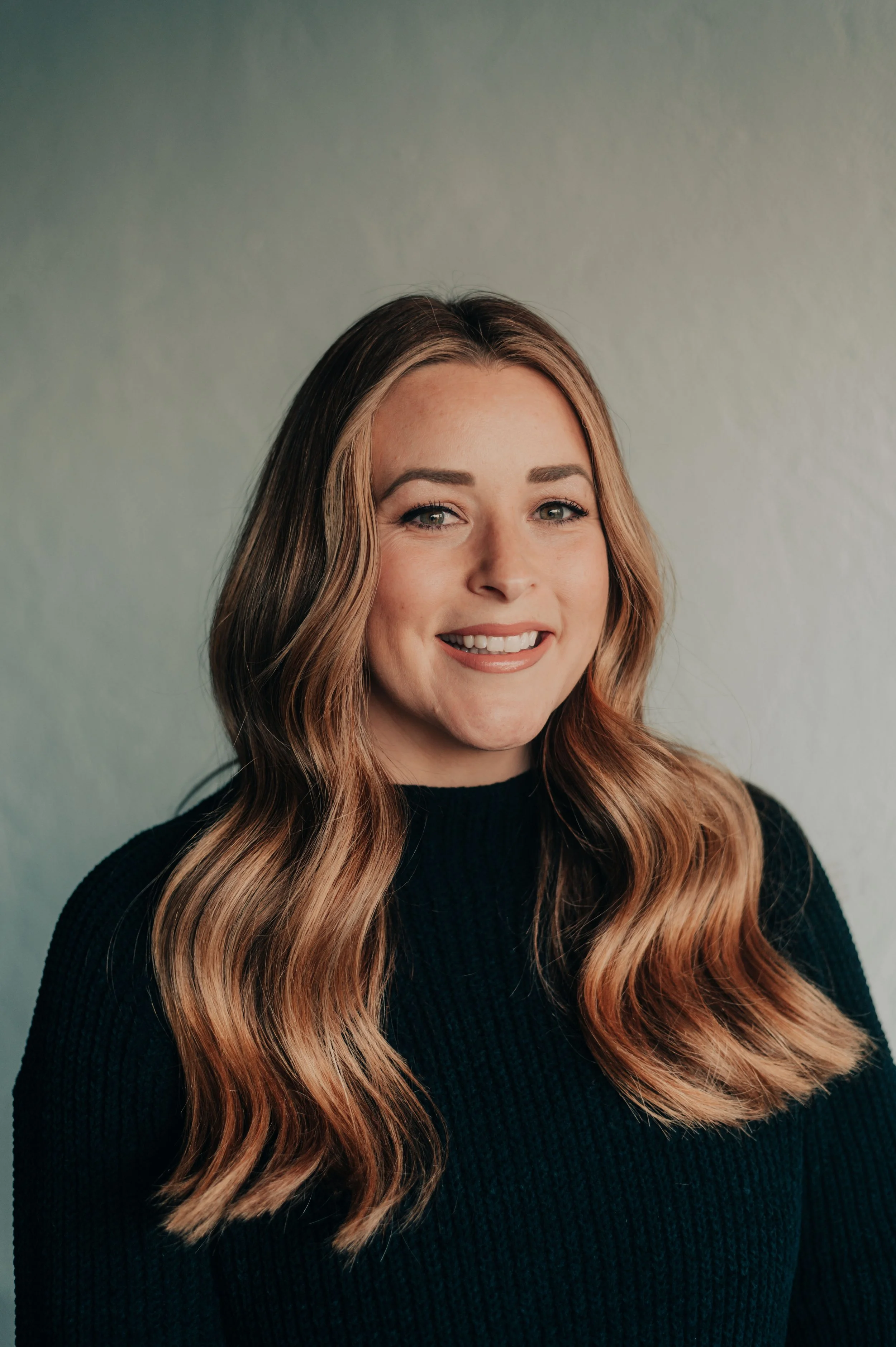 A woman with long, wavy, blonde-tinged brown hair, smiling at the camera, wearing a black sweater, against a plain neutral background.
