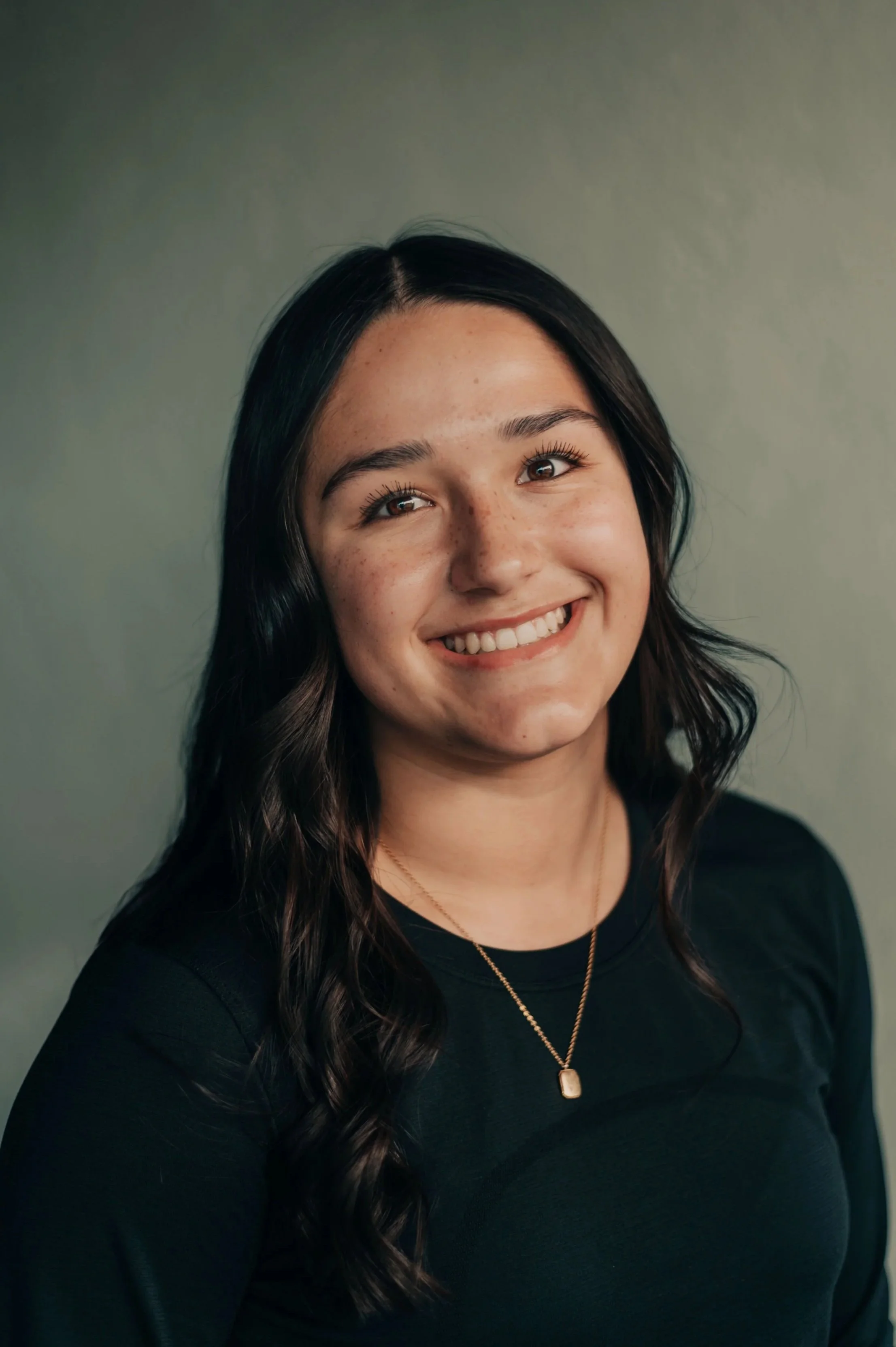 A young woman with long wavy dark hair, wearing a black top and a gold necklace with a rectangular pendant, smiling in front of a plain gray background.