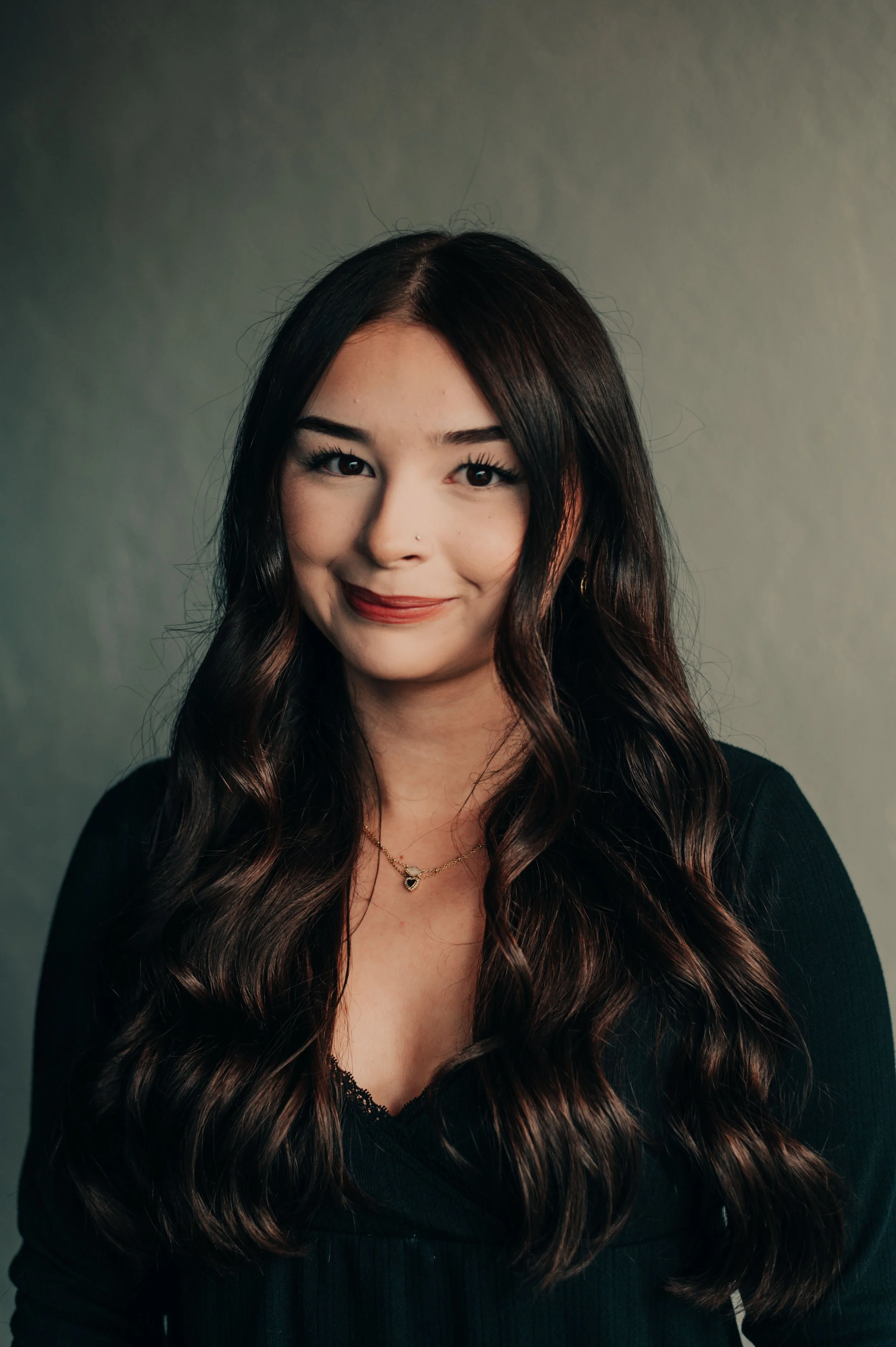 Portrait of a woman with long wavy brown hair, wearing a black top and gold necklace, smiling against a plain background.