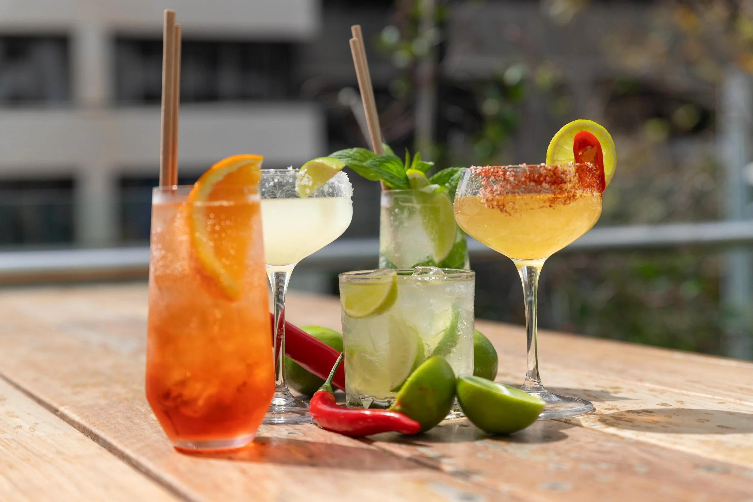 Assorted colorful cocktails with lime, lemon, and chili garnishes on a wooden table.