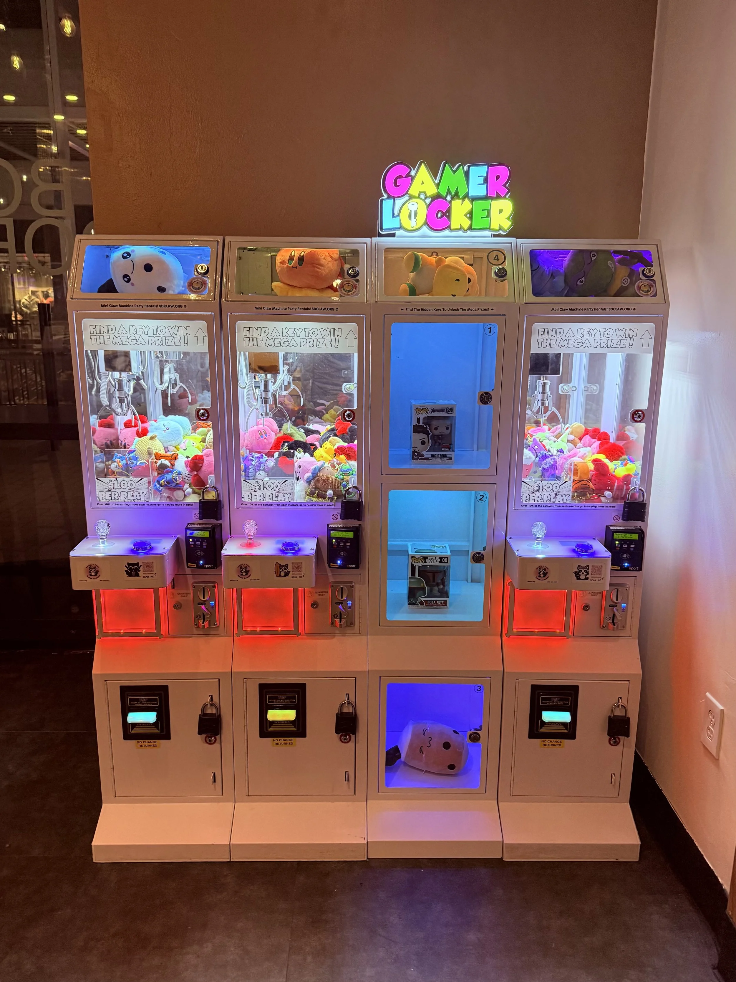 A set of four claw machine arcade games filled with plush toys, with colorful lights and a sign that reads "GAMER LOCKER" above them.