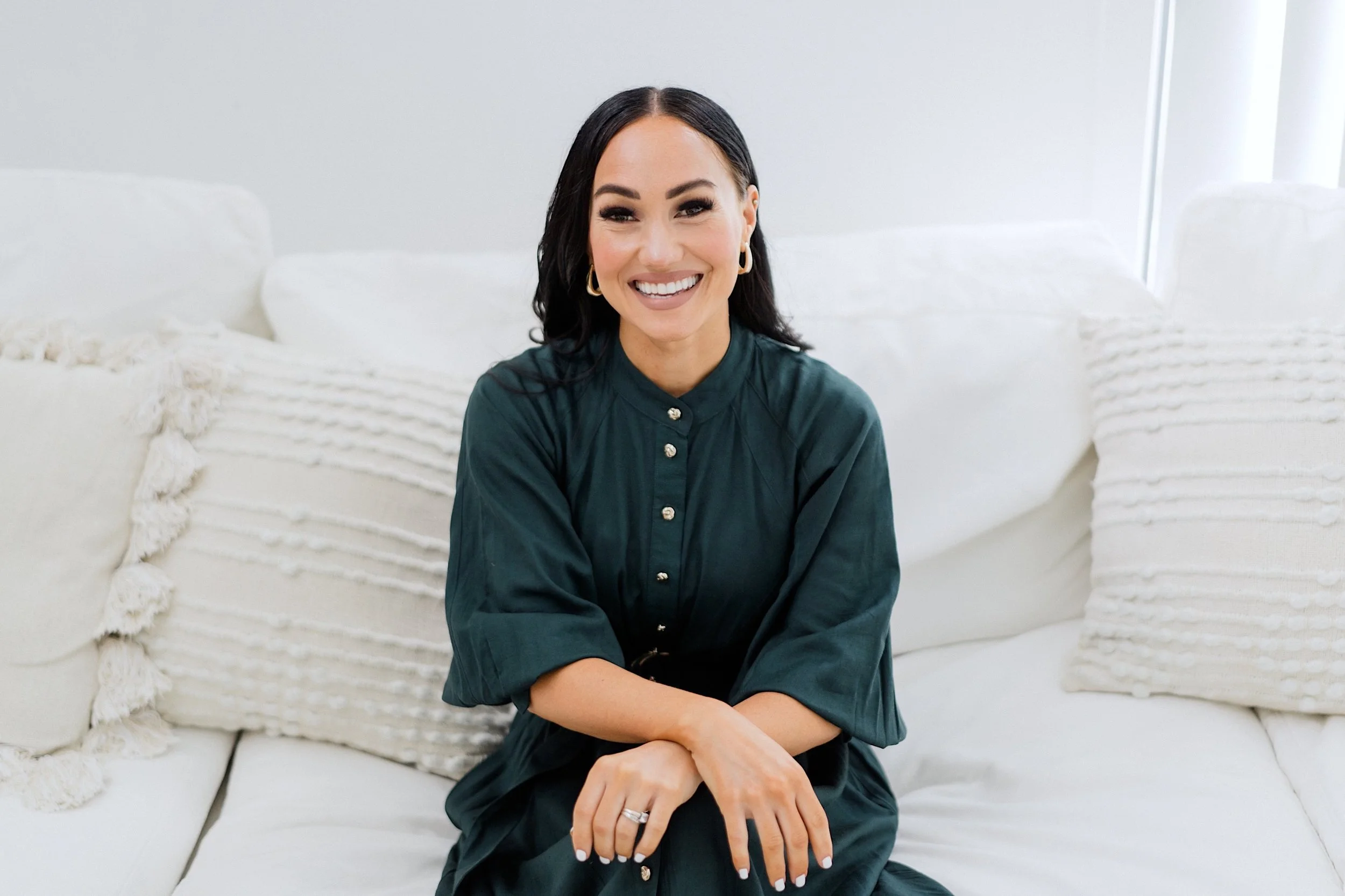 A woman with dark hair and earrings sitting on a white sofa, smiling and looking at the camera. She is wearing a dark green button-up shirt and has her hands resting on her lap.