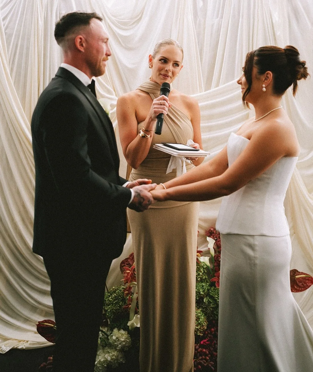 A bride and groom exchanging wedding vows, holding hands, while a bridesmaid officiates, holding a microphone and a book in front of cream-colored drapery and floral decorations.