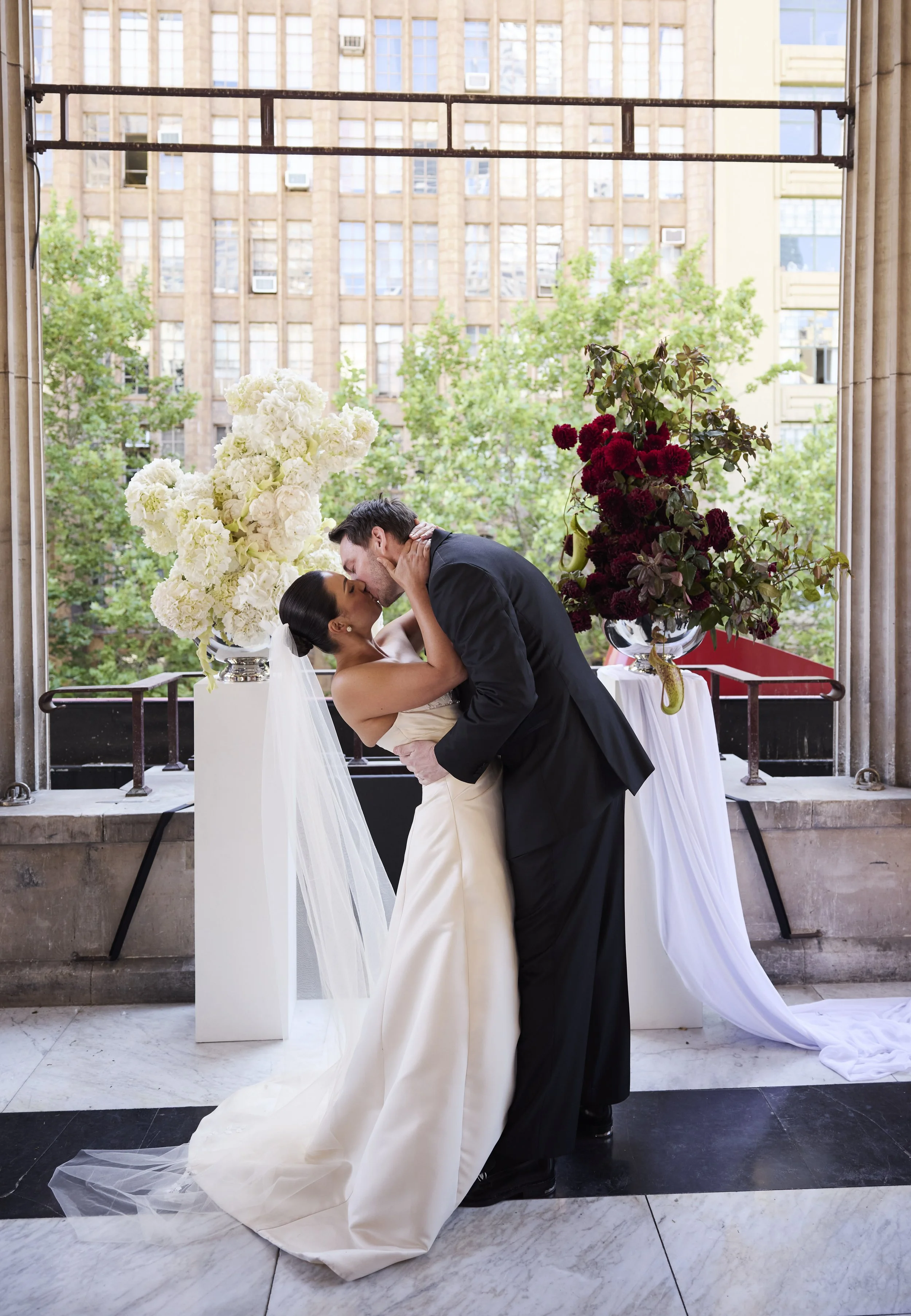 A bride and groom sharing a kiss during wedding vows, with floral arrangements and a cityscape background.