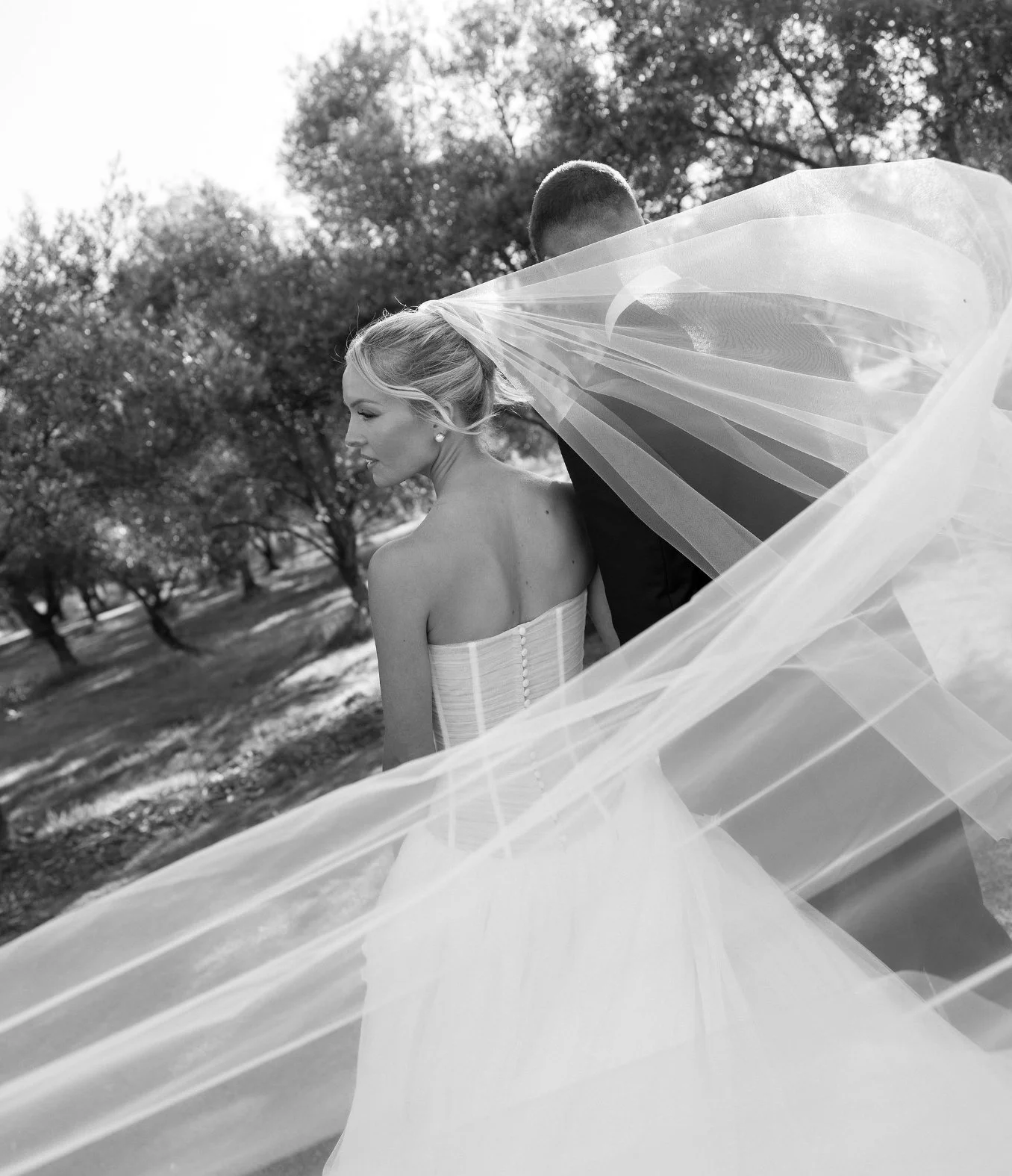 Black and white photo of a bride in a strapless wedding dress with her hair styled in loose waves, standing outdoors with trees in the background. A groom, partially obscured by her veil, stands beside her.