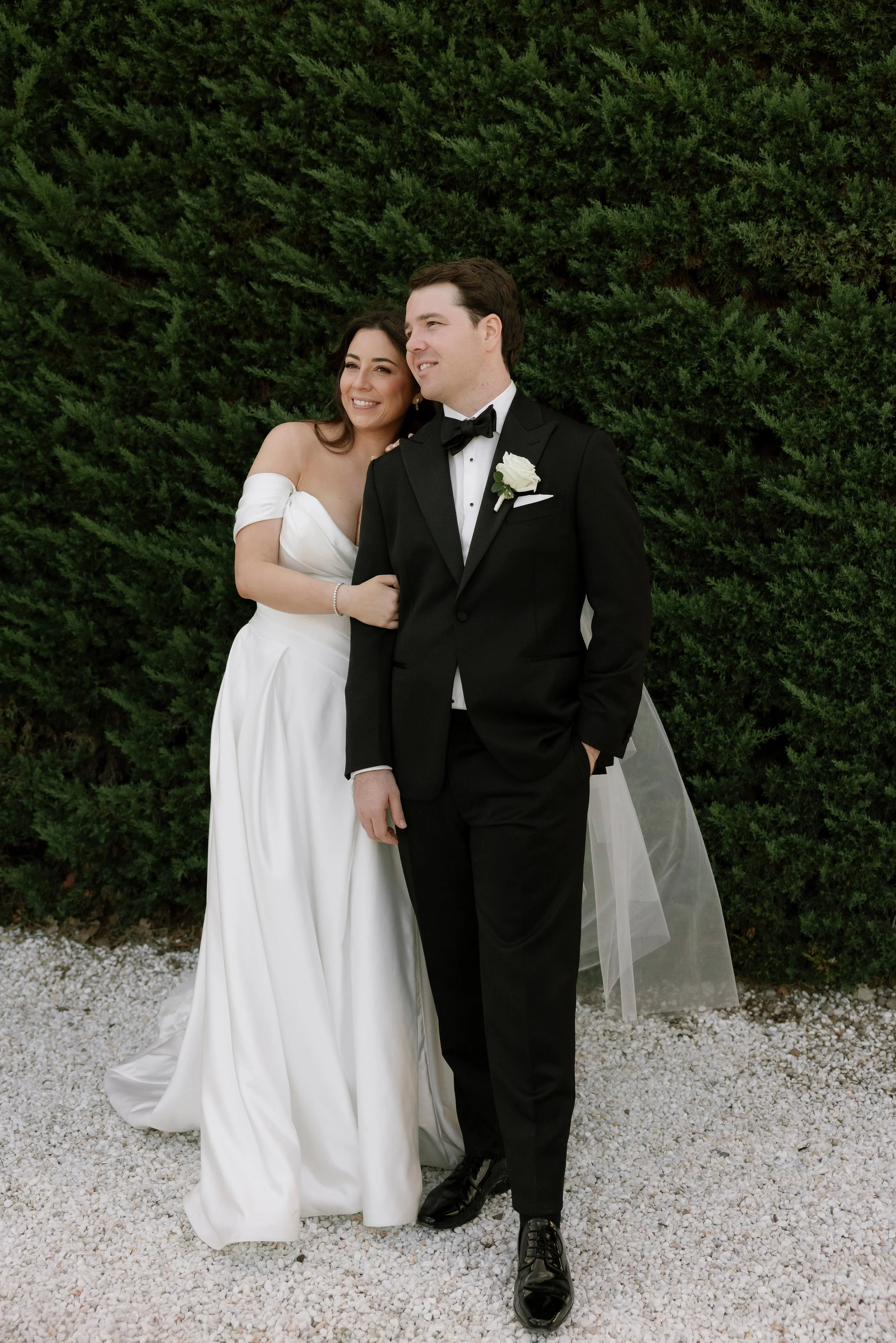 A bride and groom standing together outdoors on a gravel surface with green shrubbery in the background, smiling and dressed in wedding attire.