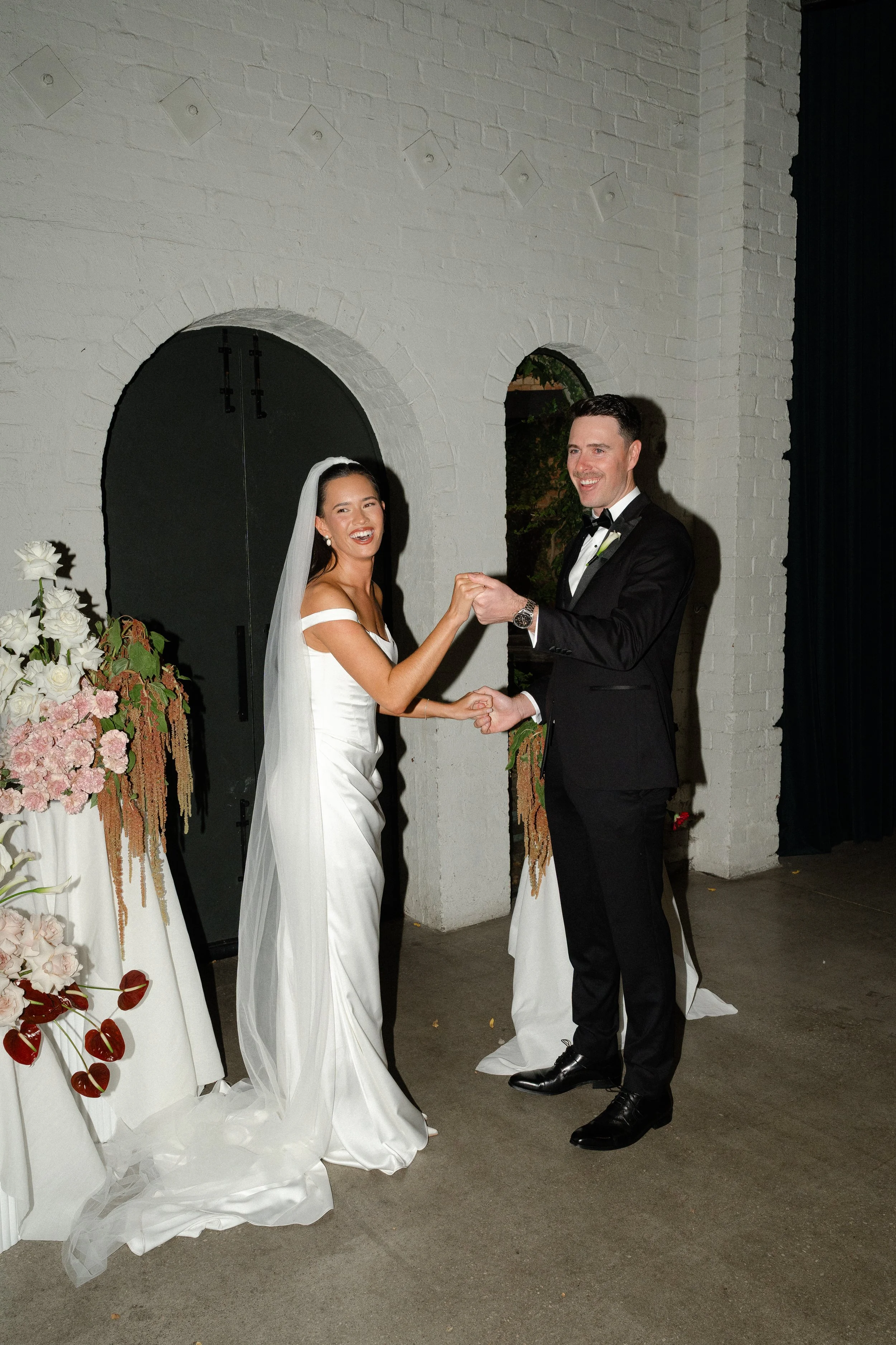 A bride in a white wedding gown and veil holding hands with a groom in a black tuxedo, smiling and dancing at their wedding reception with floral decorations nearby.