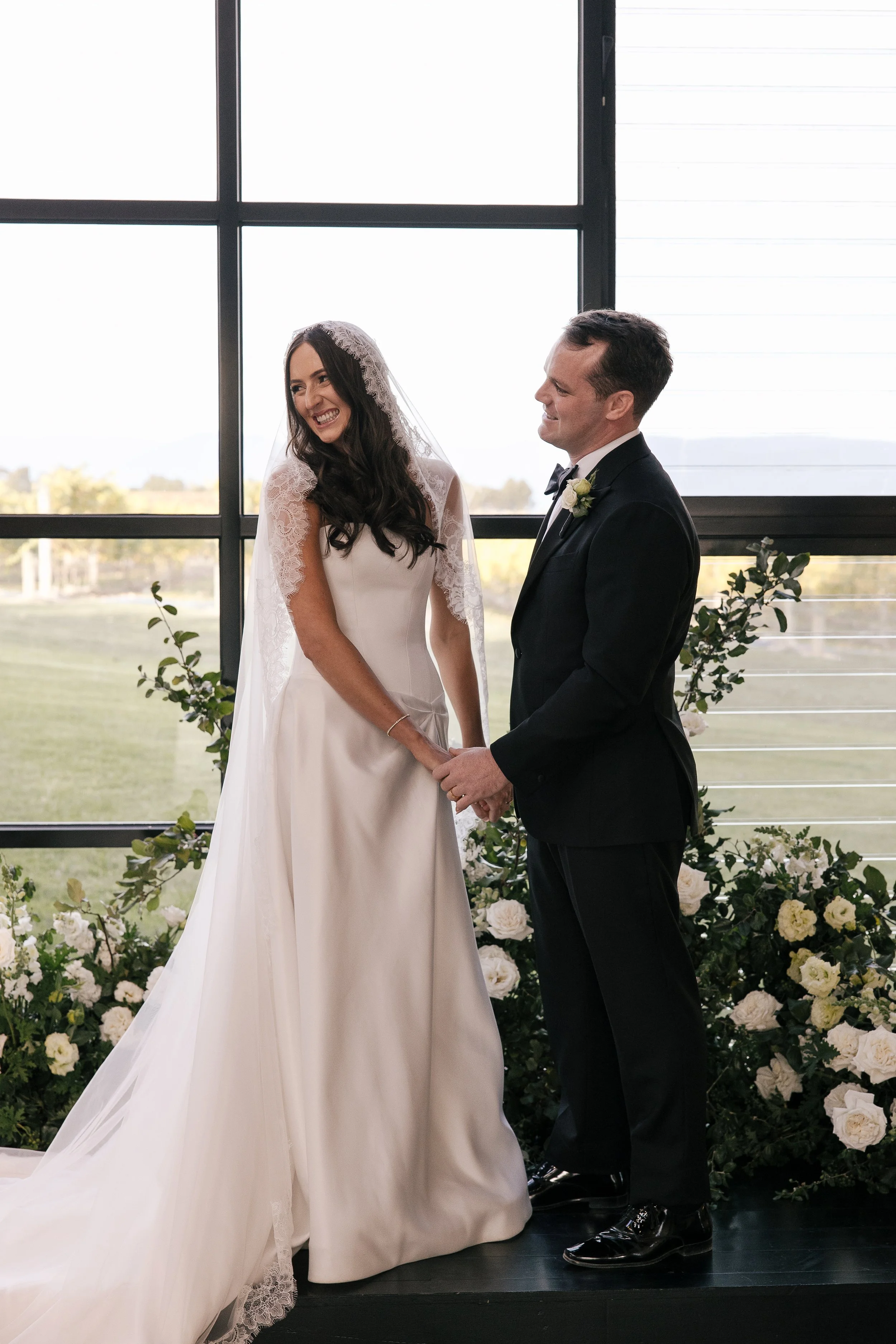 A bride and groom are exchanging vows at an indoor wedding ceremony with large windows and a view of the outdoors, surrounded by floral decorations.