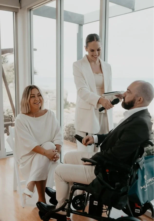 A woman in a white suit conducts an interview with a man in a wheelchair in a bright room with large windows.