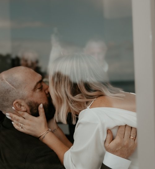 Close-up of a man and woman sharing a kiss behind a glass window, with reflections visible in the glass.